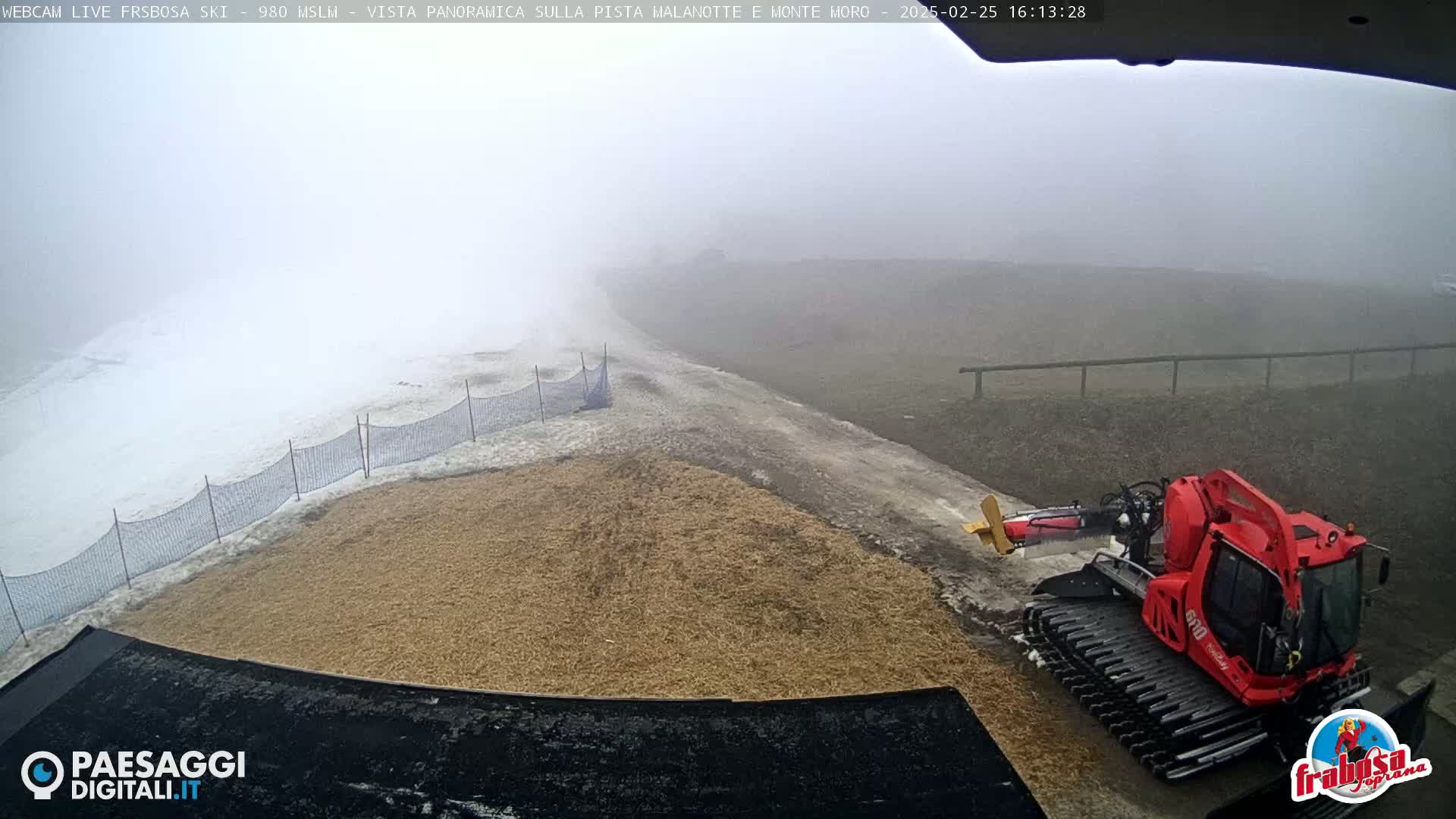 A snowcat sits on a foggy, snow-covered slope next to a patch of tan ground, with a snow fence in the background.