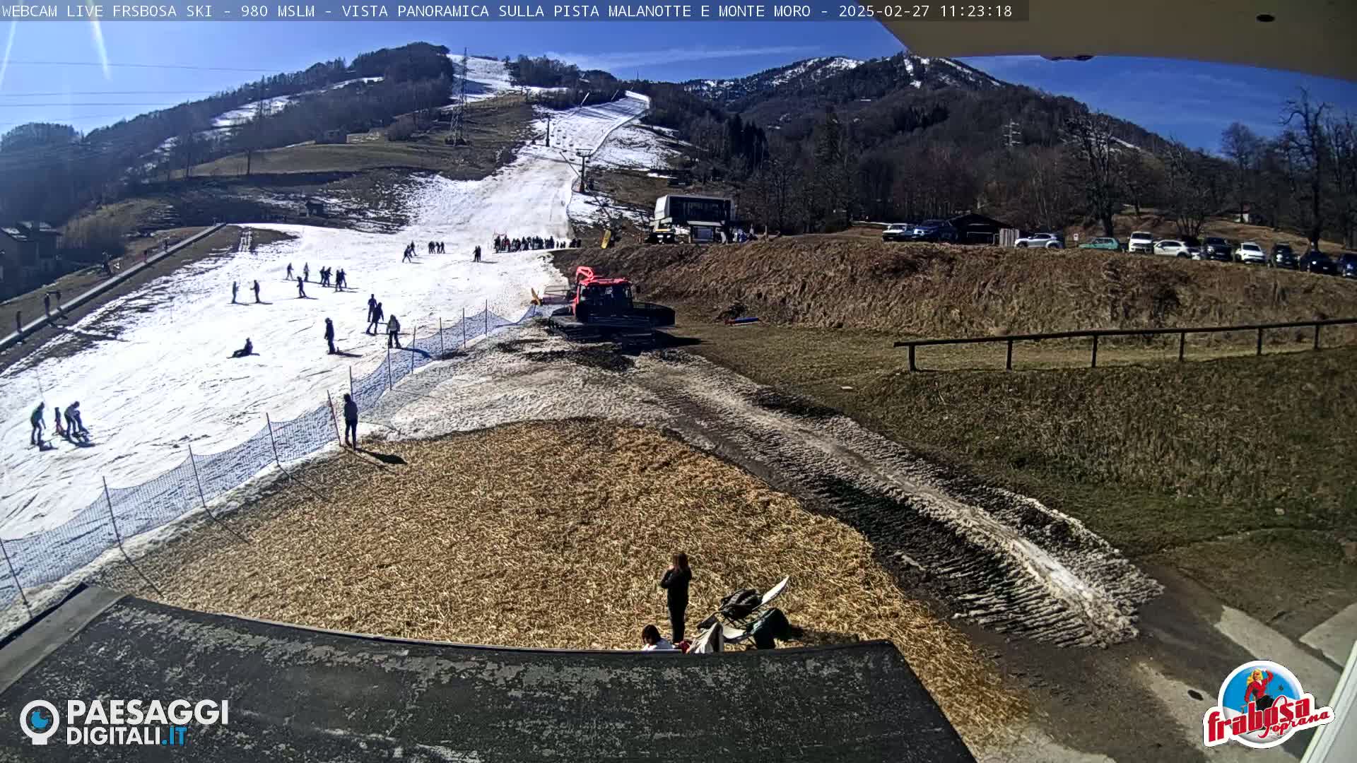 A sunny day at a ski resort shows a snow-covered slope with skiers and snowboarders, a snowcat, and several people near a tan-colored area at the bottom of the slope.