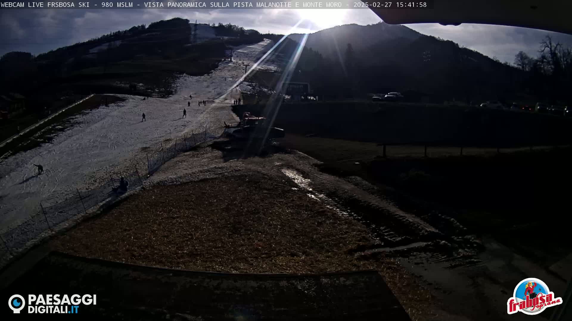 A sunny, snow-covered ski slope with a snowcat parked at its base, several skiers on the slope, and a muddy area below.