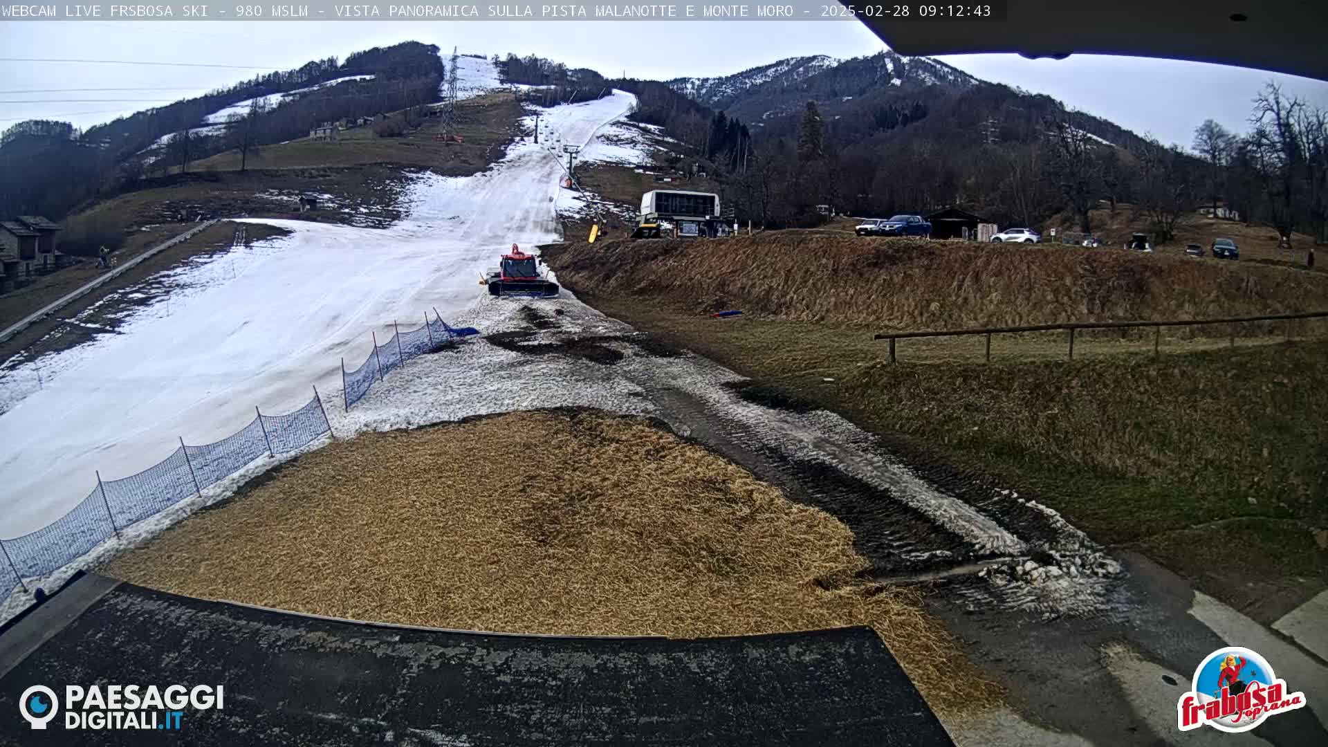 A snow groomer is parked near a snow-covered ski slope on a partly cloudy day, with a pile of hay or straw nearby.