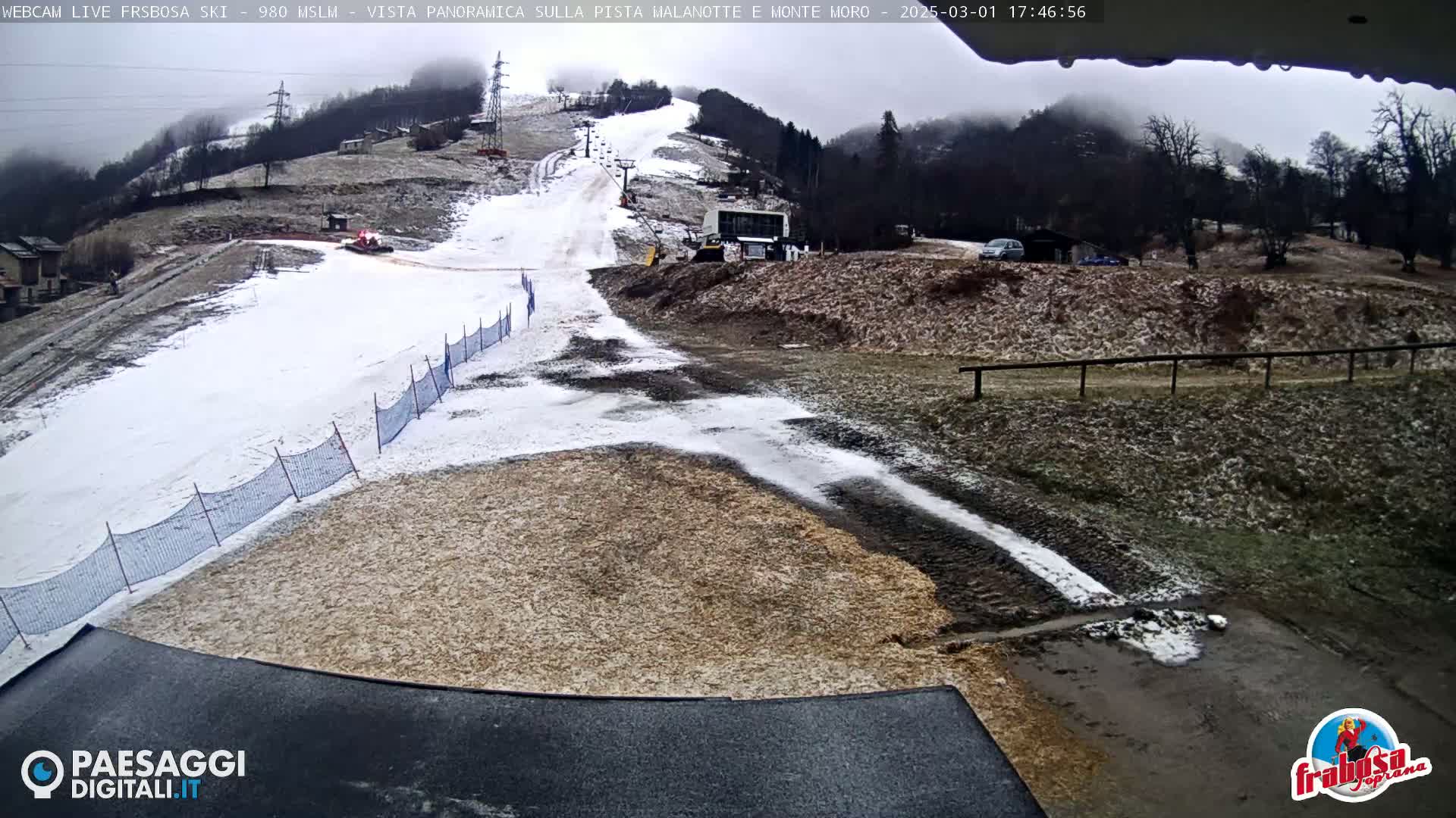A snow-covered ski slope with a snowcat and a chairlift visible in the overcast weather.