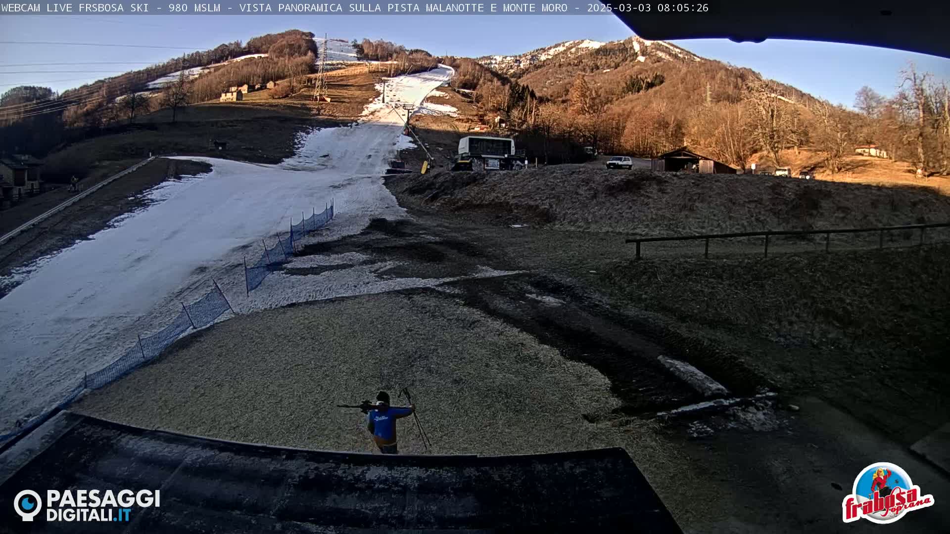 A person with skis stands in a snow-covered ski area, with a mountainside visible in the background under a sunny sky.