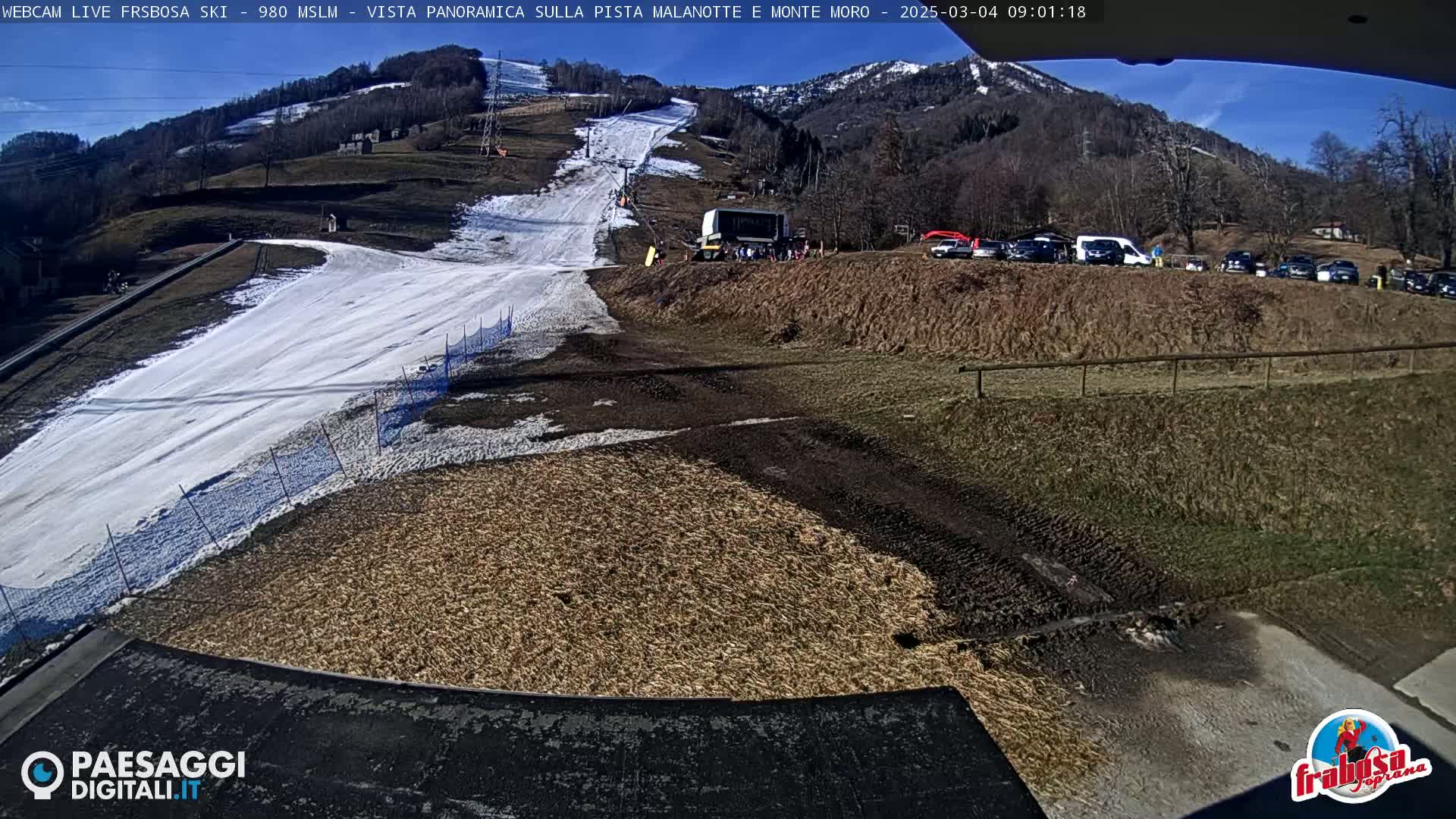 A snow-covered ski slope on a sunny day is partially visible, with a muddy area below, several vehicles parked nearby, and a mountain in the background.