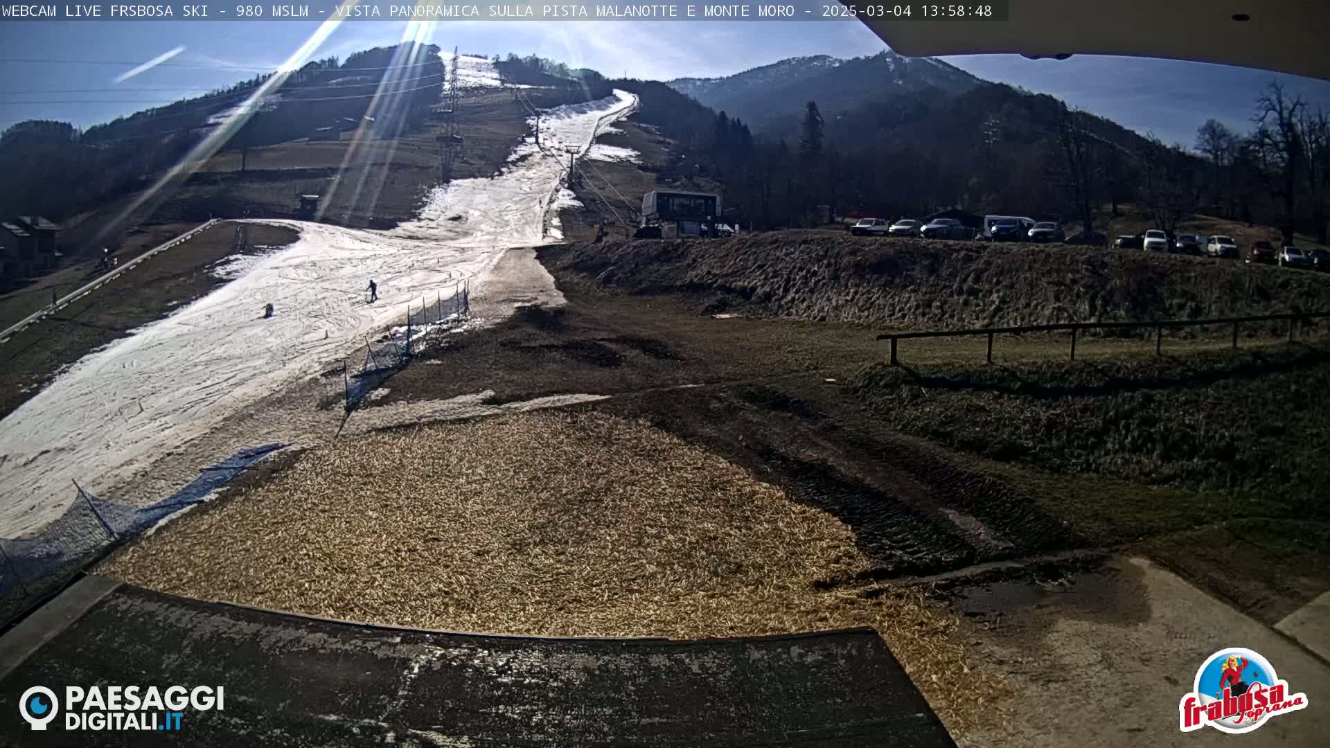 A sunny day at a ski slope shows a partially snow-covered hill with a few skiers, a snow-grooming machine, and a parking lot full of cars.