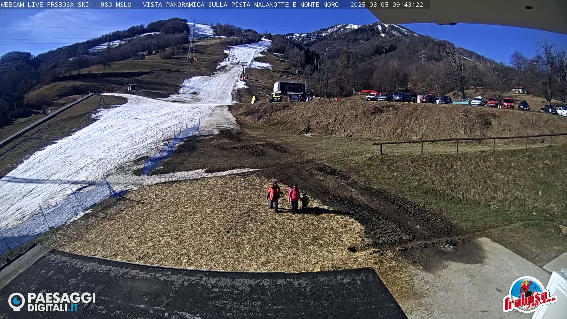 A sunny, snow-covered ski slope is visible from a high vantage point, with a few people standing near a lower, dirt section of the slope and several cars parked on a hillside in the background.