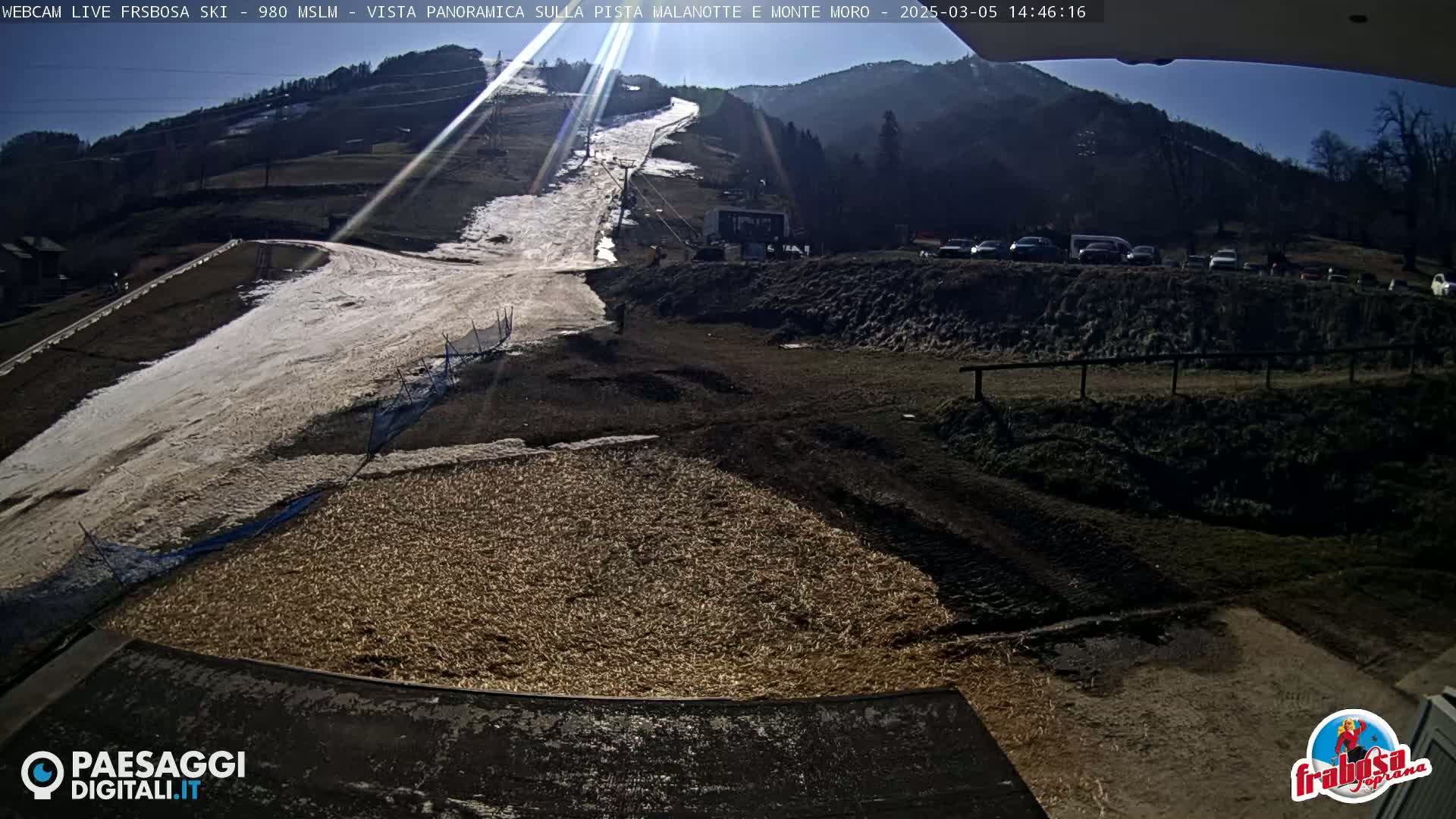 A sunny view of a snow-covered ski slope with a parking lot and surrounding hills.