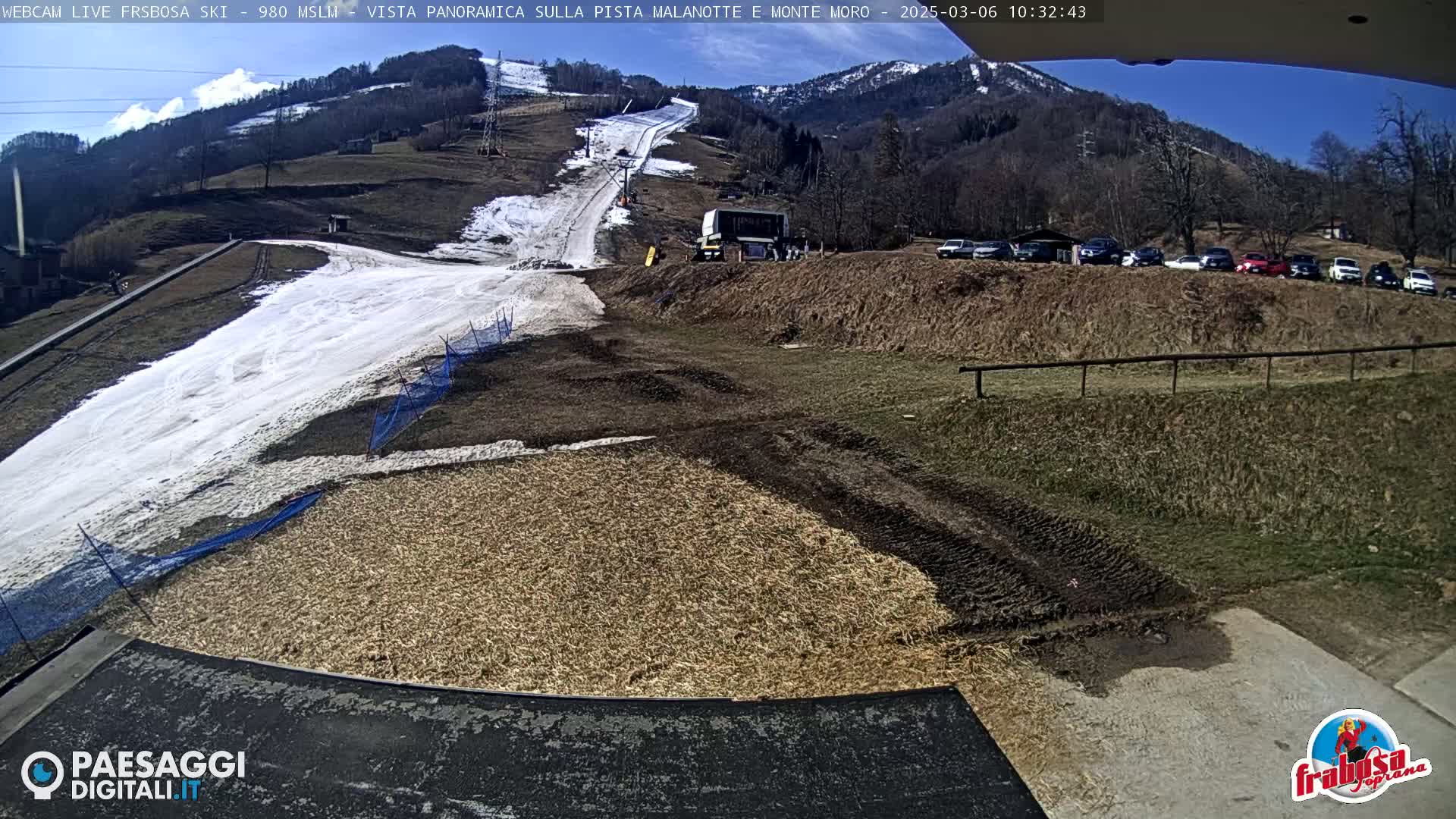A sunny view of a ski slope with patchy snow, a snow-grooming machine, and a parking lot full of cars, all situated on a hillside with brown grass and bare trees.