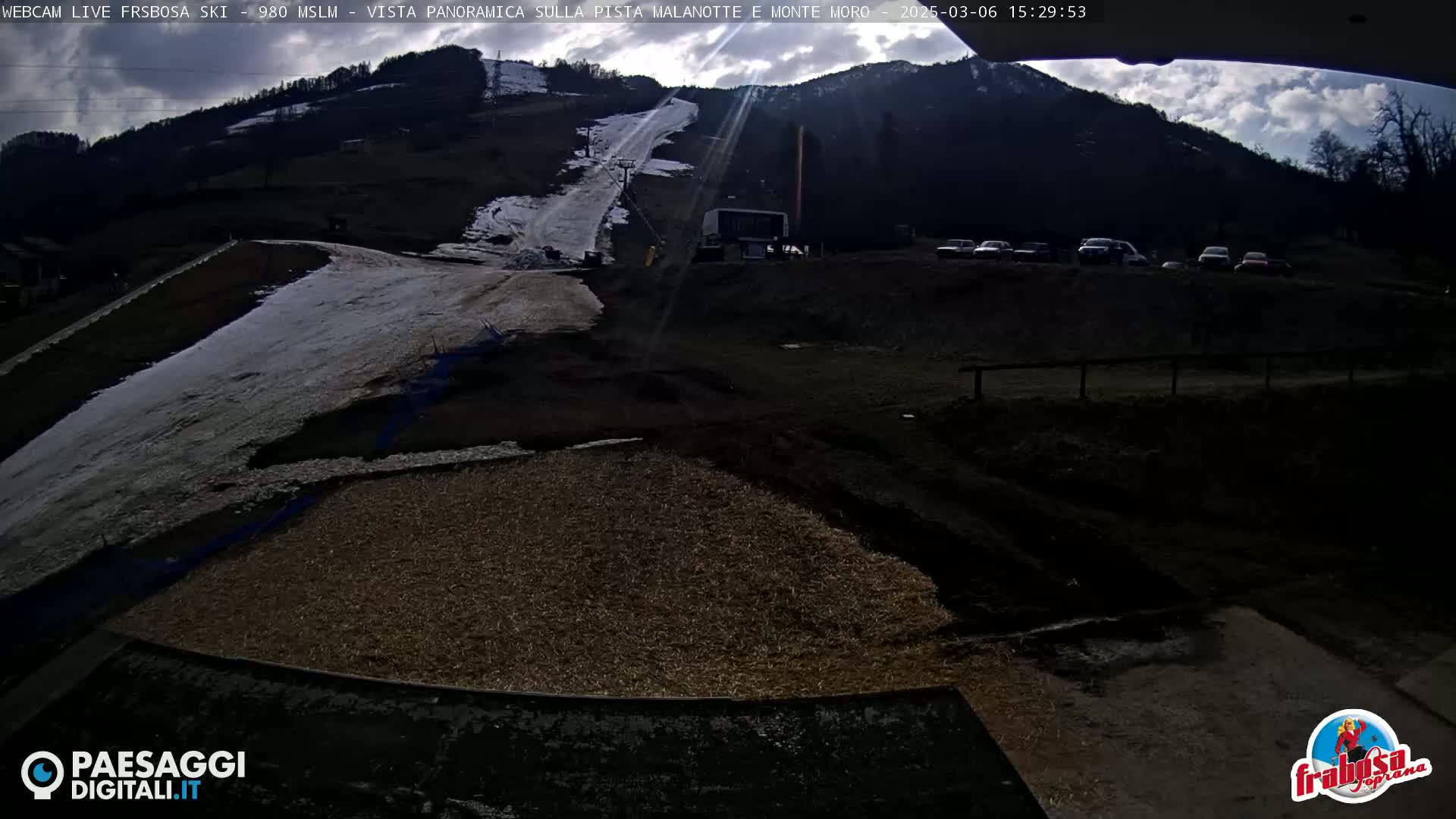 A mostly sunny day at a ski resort shows a partially snow-covered ski slope, several parked cars, and a mostly bare brown ground in the foreground.
