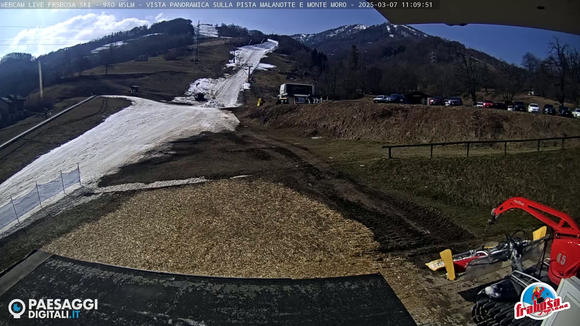 A sunny day at a ski resort shows a snow-covered slope with a ski lift, a parking lot full of cars, and snow-grooming equipment near the bottom of the hill.