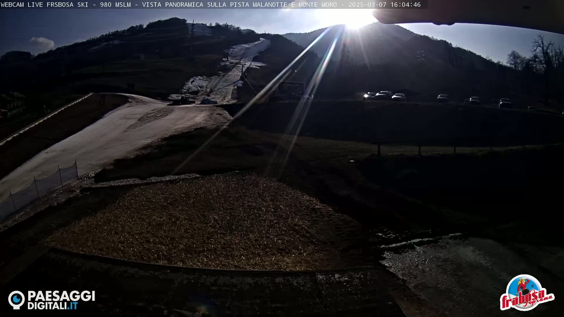 A sunny, outdoor view of a ski slope with several cars parked nearby and a large area of brown ground in the foreground.