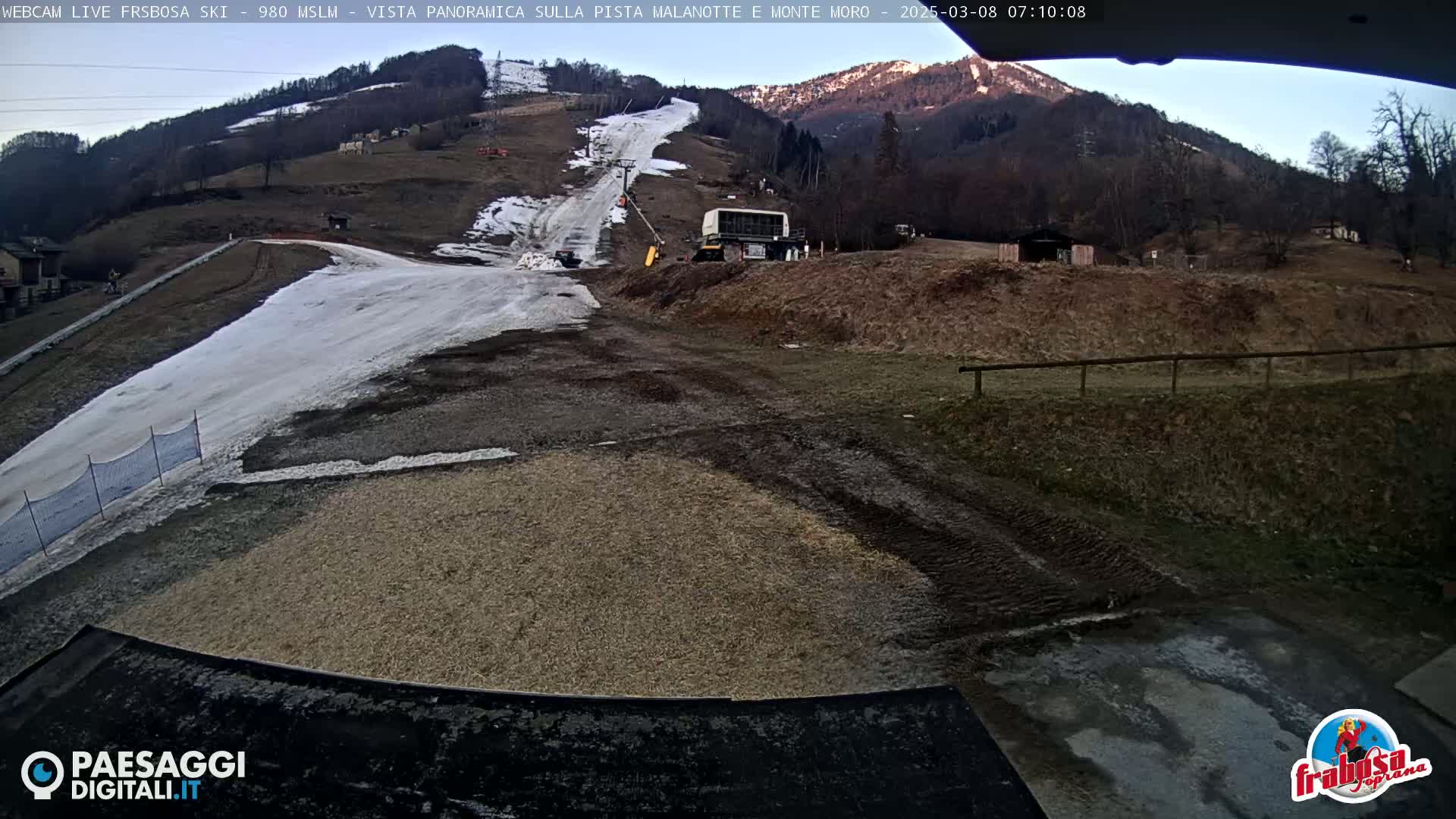 A ski slope with patches of snow and brown ground, a snow-grooming vehicle, and a mountain background under a mostly clear sky.