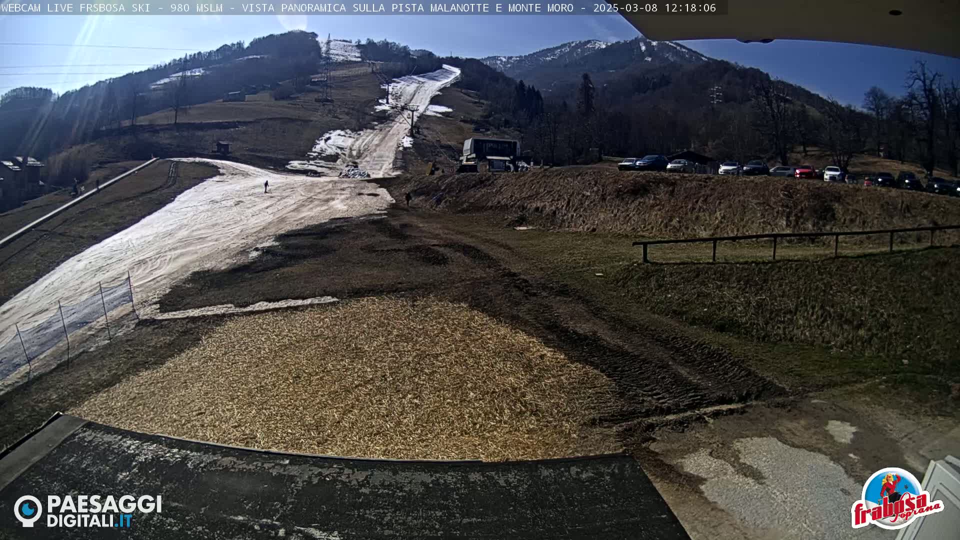 A sunny, snow-covered ski slope with a partially muddy base, a few parked cars, and a mountain in the background.