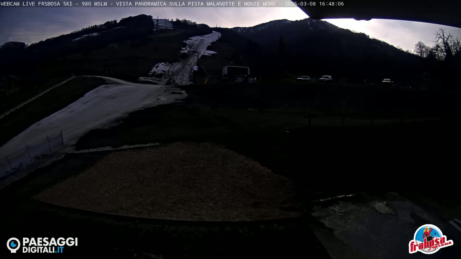 A mostly dark, overcast scene shows a snow-covered ski slope on a mountainside, a road leading to it, and a few vehicles in a parking area.