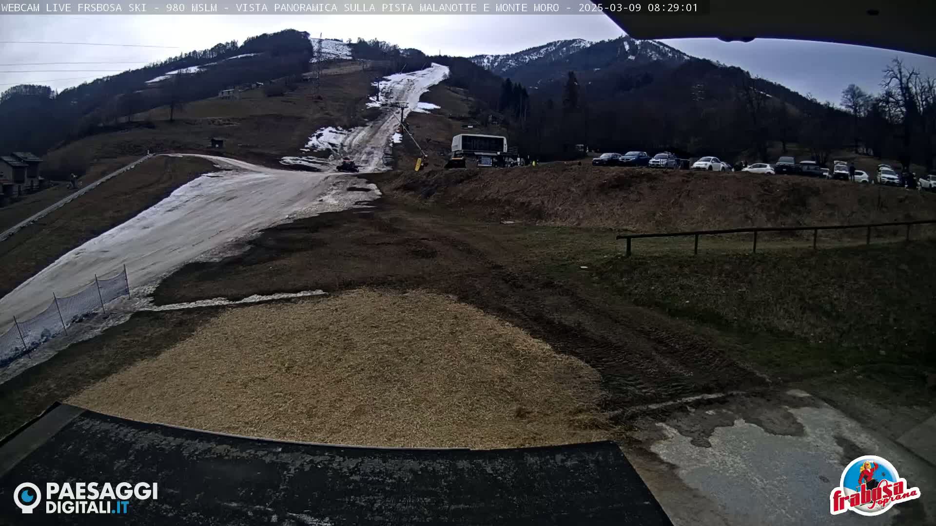 A mostly cloudy day reveals a ski slope with patchy snow, a snow-grooming machine, and several cars parked in a lot below a mountain.