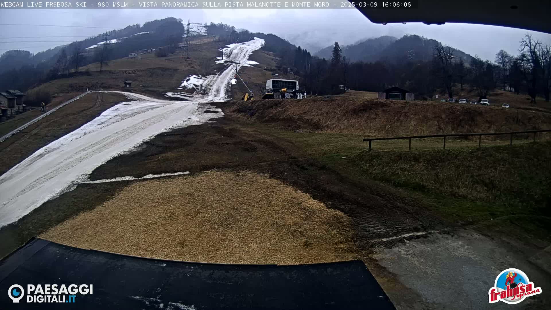 A snow-covered ski slope with a snowcat and some vehicles parked near it under an overcast sky.