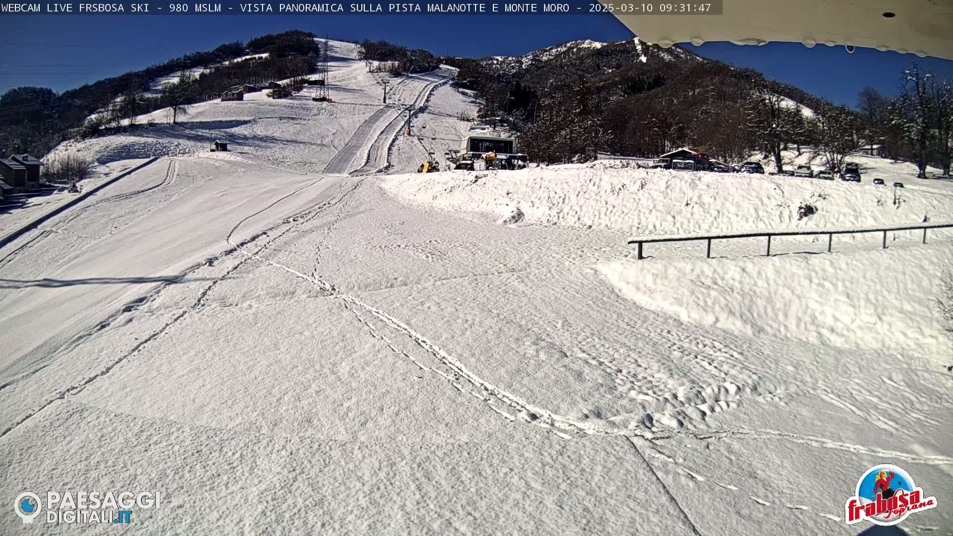 A snow-covered ski slope with a ski lift and several buildings is visible under a sunny sky.