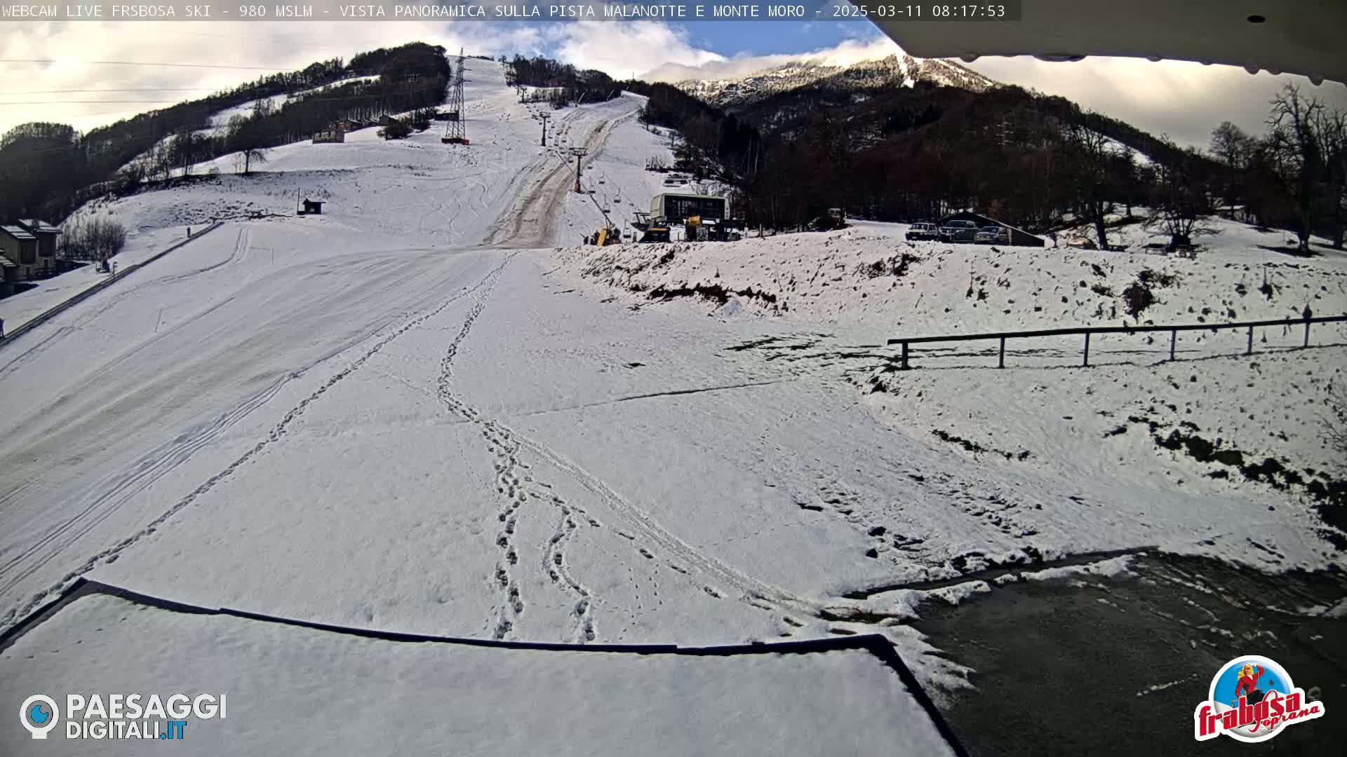A snow-covered ski slope with a ski lift and footprints in the snow, under a partly cloudy sky.