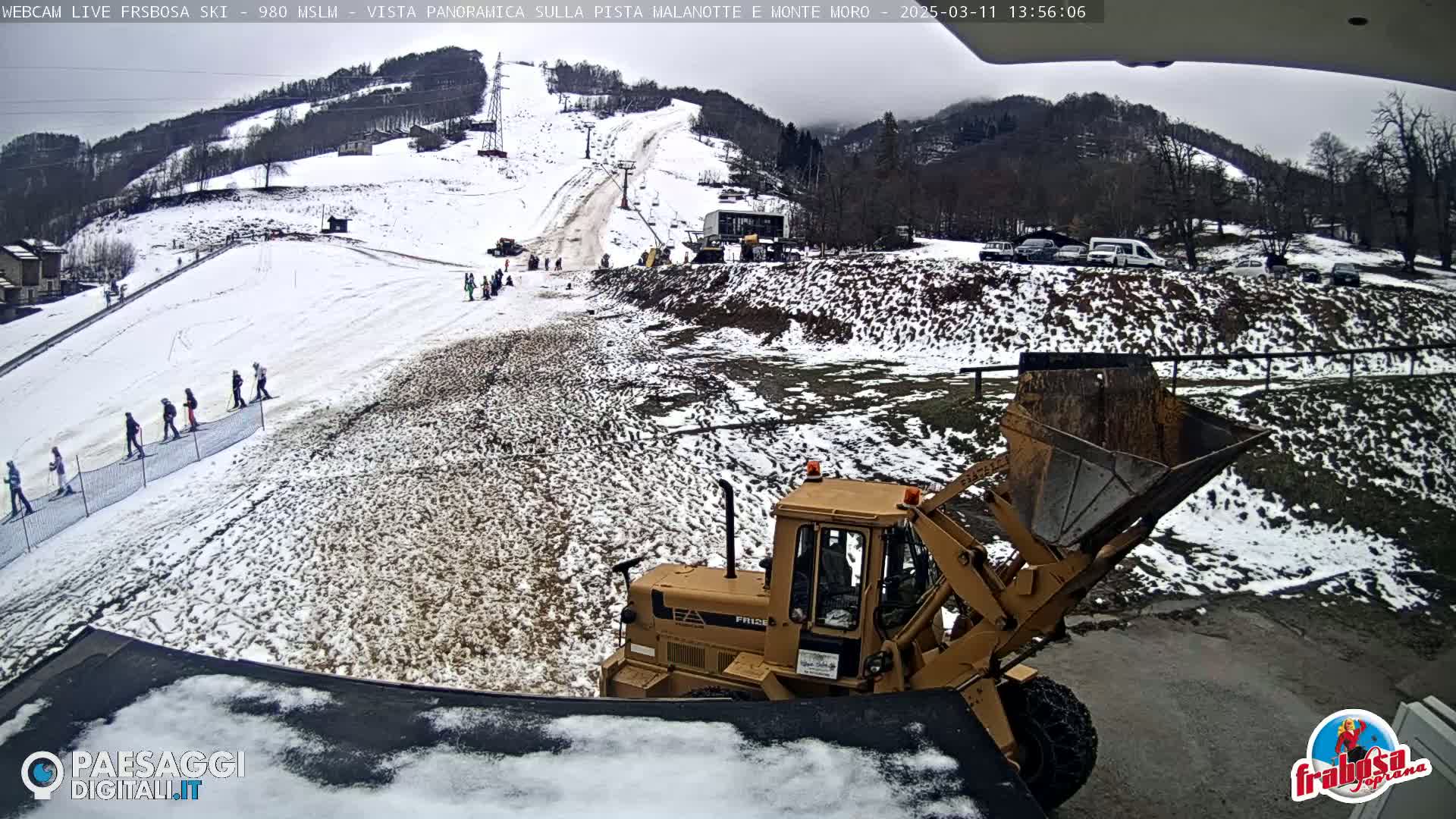A snow-covered ski slope with several skiers, a snowplow, and a few parked vehicles is visible under an overcast sky.