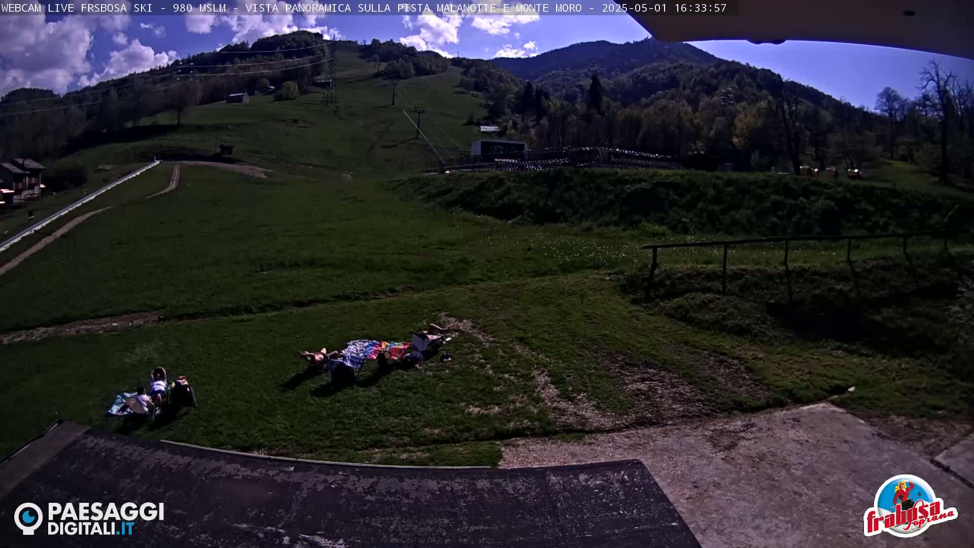 A sunny outdoor scene shows a grassy hillside with a few people relaxing on blankets near a paved area, overlooking a mountain in the distance.