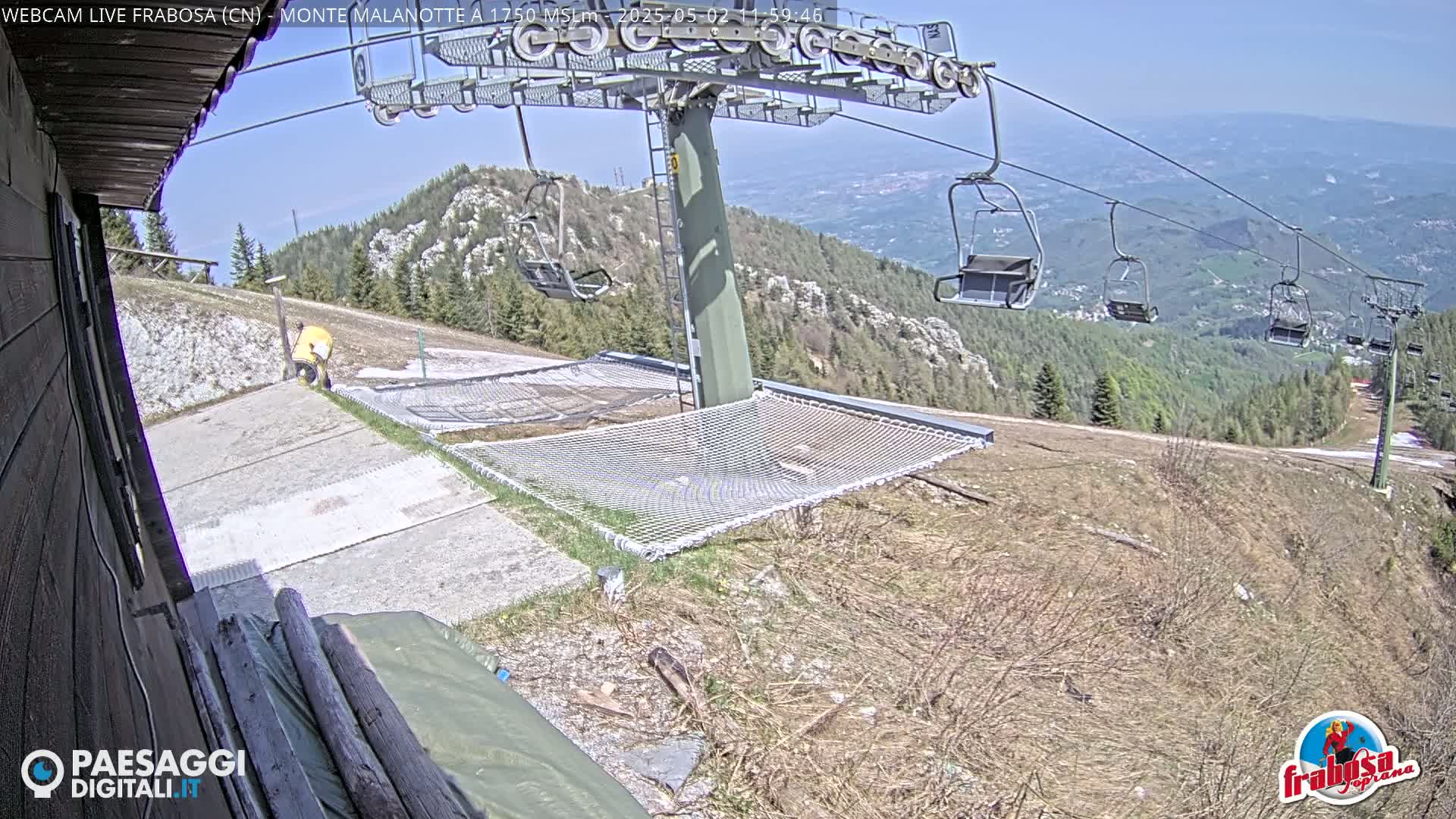A person is seen near a ski lift and safety net on a mountainside under a clear, sunny sky, with distant valleys visible.