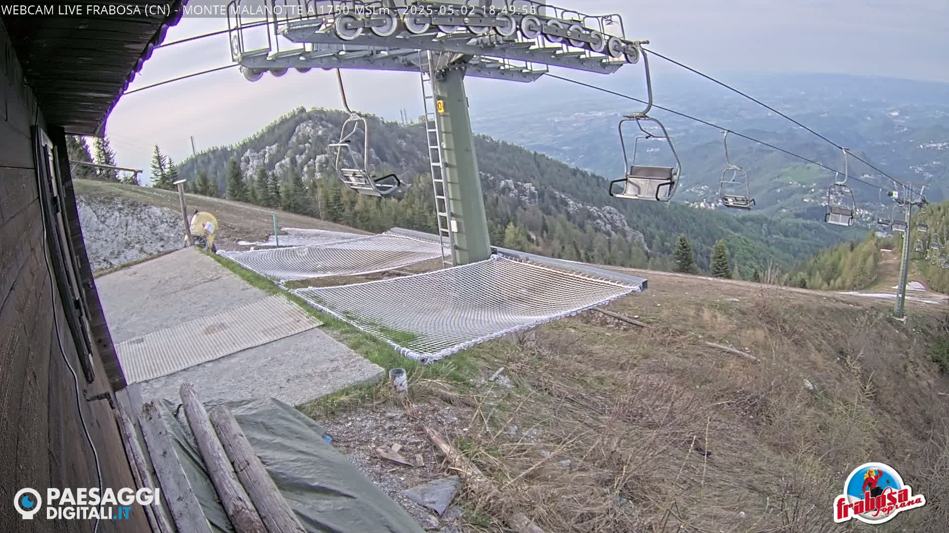 A mountaintop ski lift with empty chairs overlooks a valley under partly cloudy skies, with several safety nets visible at the base of the lift.