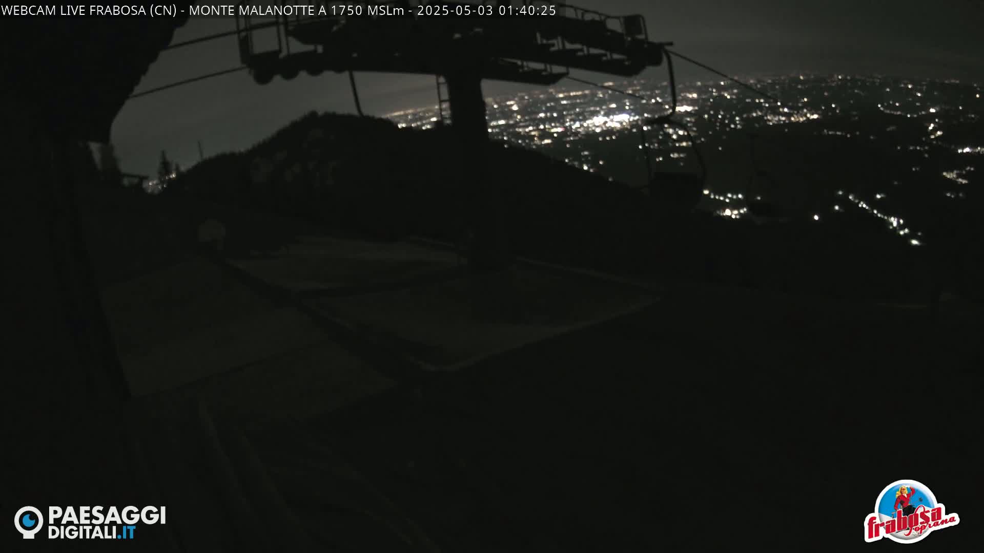 A nighttime view from a mountaintop shows a chairlift and a distant city illuminated below.