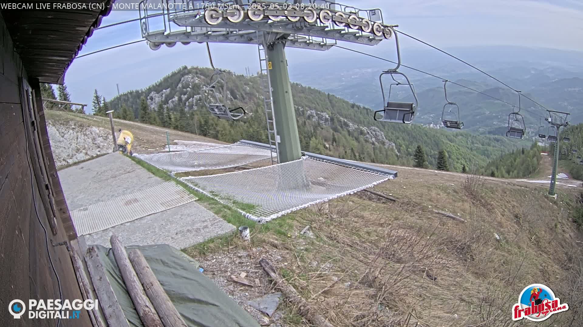 A ski lift with empty chairs overlooks a grassy mountainside with safety nets and a person in yellow near a building under a mostly clear sky.