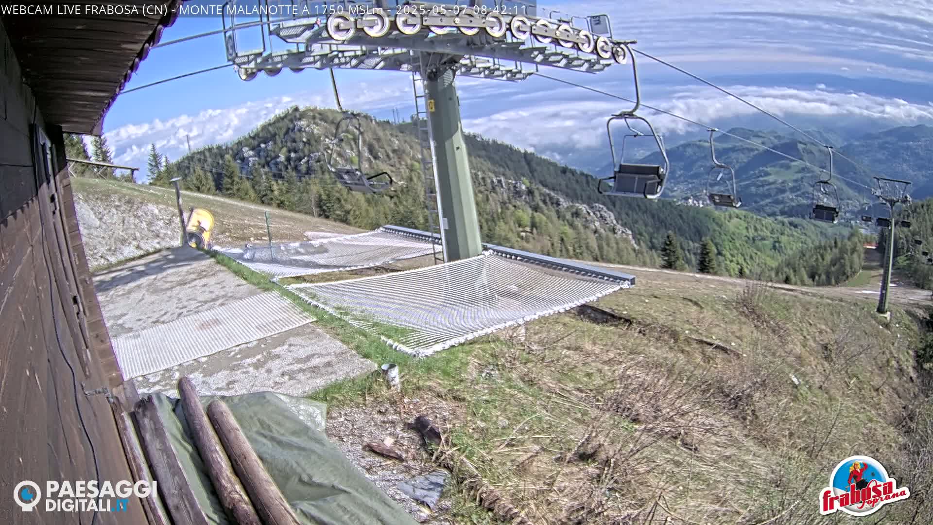 A partly cloudy day reveals a mountaintop scene with a chairlift, several empty chairlift seats, and two empty mesh hammocks near a gravel area.