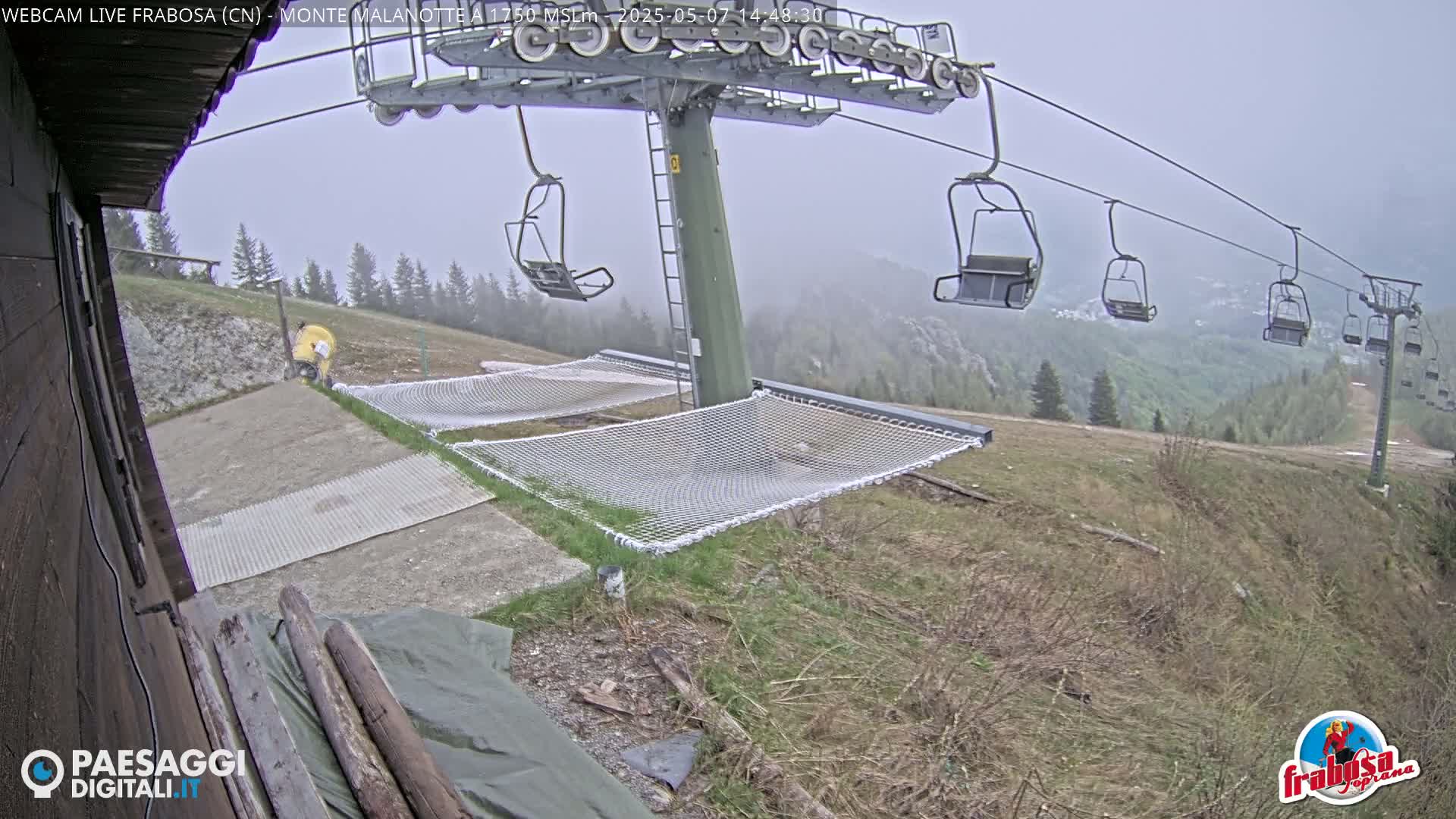 A chairlift with empty chairs overlooks a grassy hill with two empty safety nets in foggy conditions.