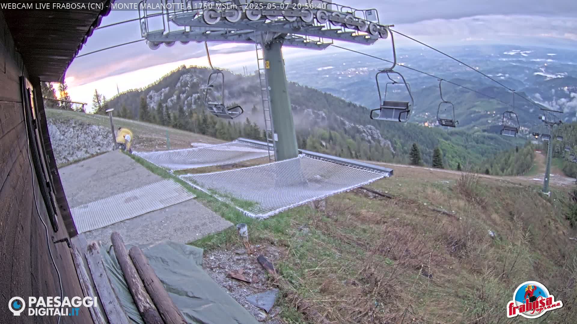 A mountaintop view at dusk shows an empty chairlift with several hammocks near a building, overlooking a valley.