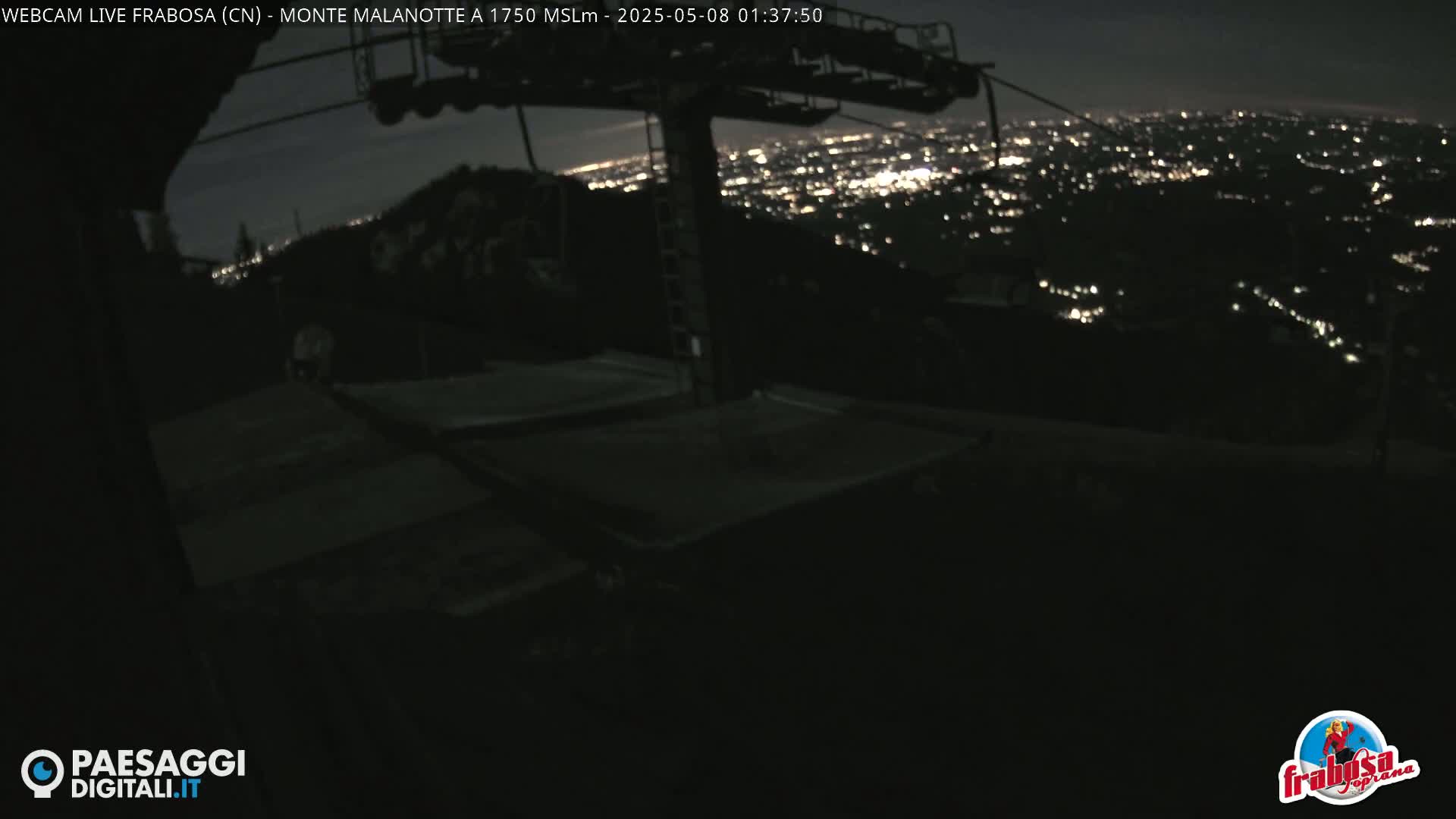 A nighttime view from a mountaintop shows a chairlift and a distant city glowing with lights.