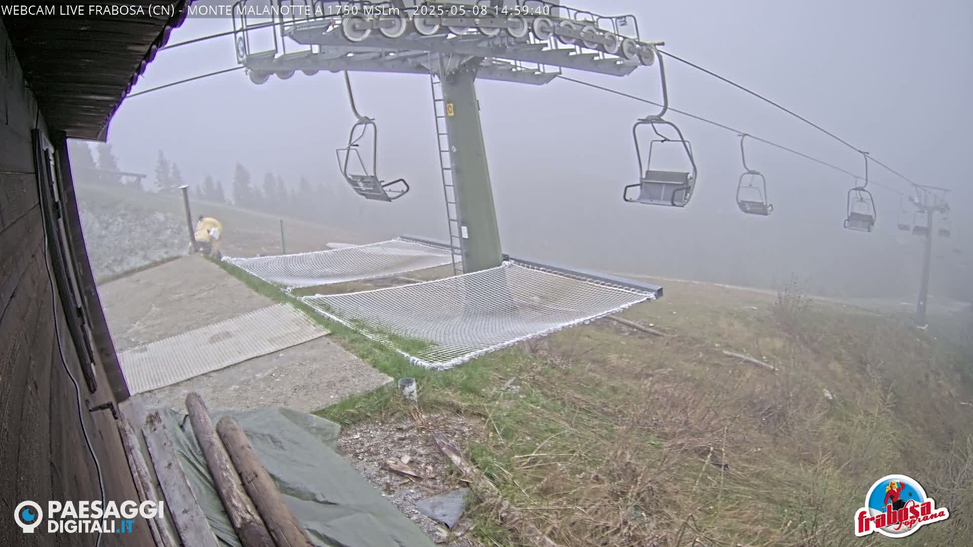 A foggy mountaintop scene shows a chairlift with empty chairs, safety netting, and a person in yellow clothing near the base.