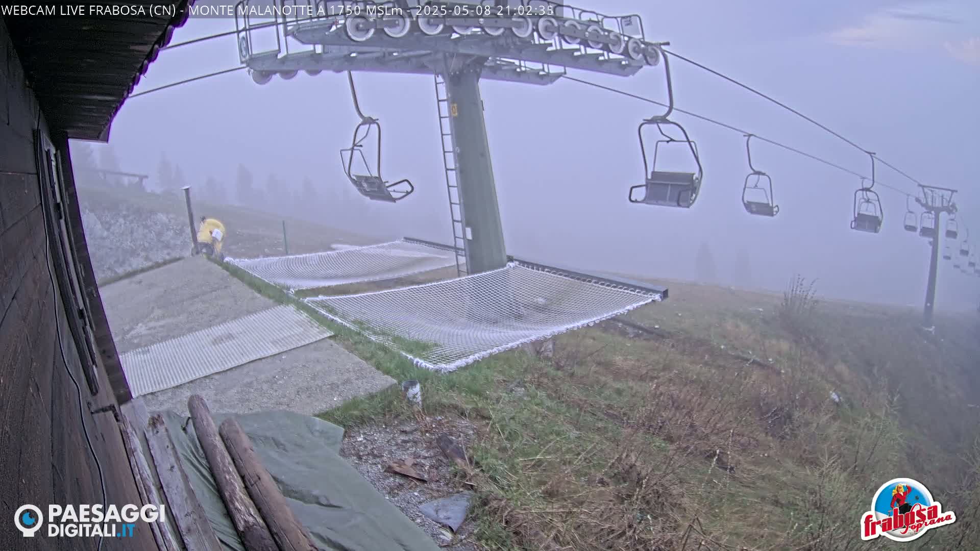 A foggy mountaintop scene shows a chairlift with empty seats, two mesh hammocks, and a person in yellow standing near a metal grating.