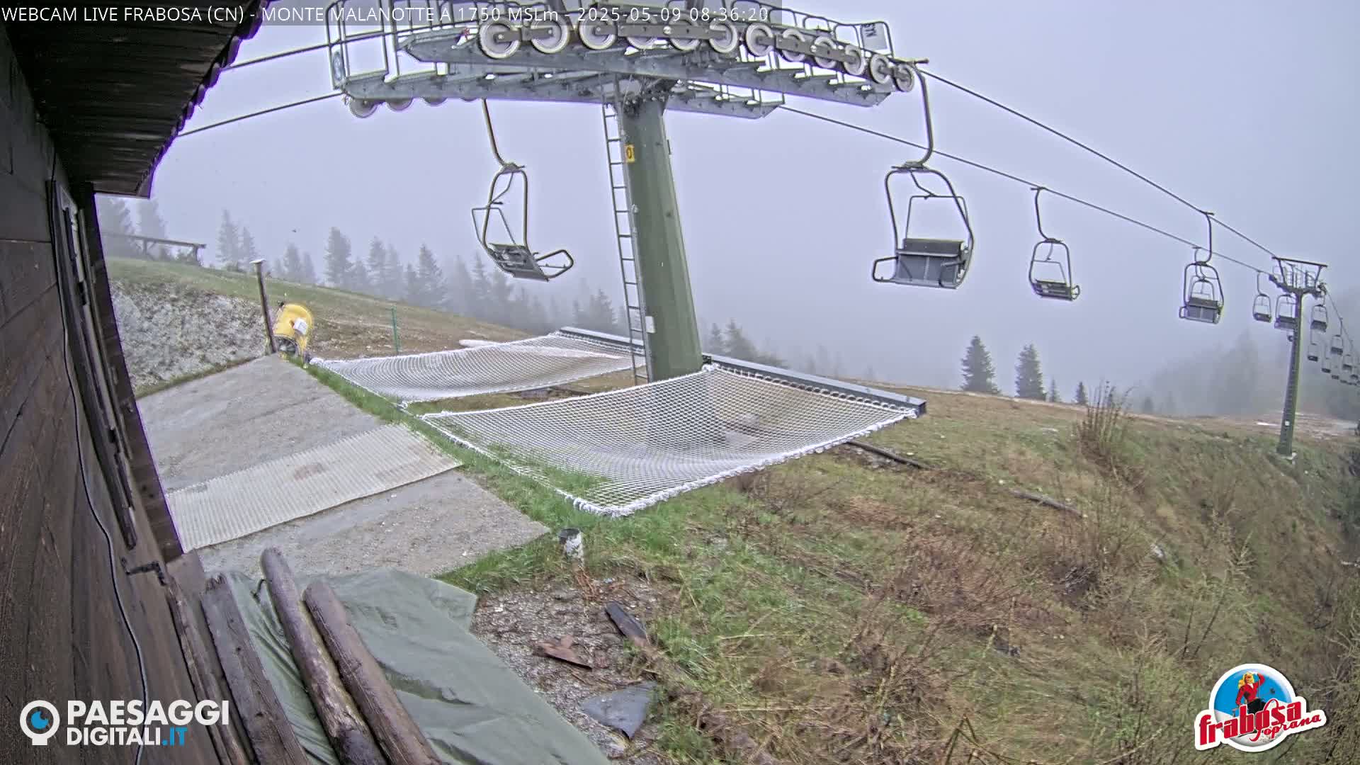 A chairlift with empty chairs ascends a foggy mountainside where safety nets are set up near a concrete unloading area.
