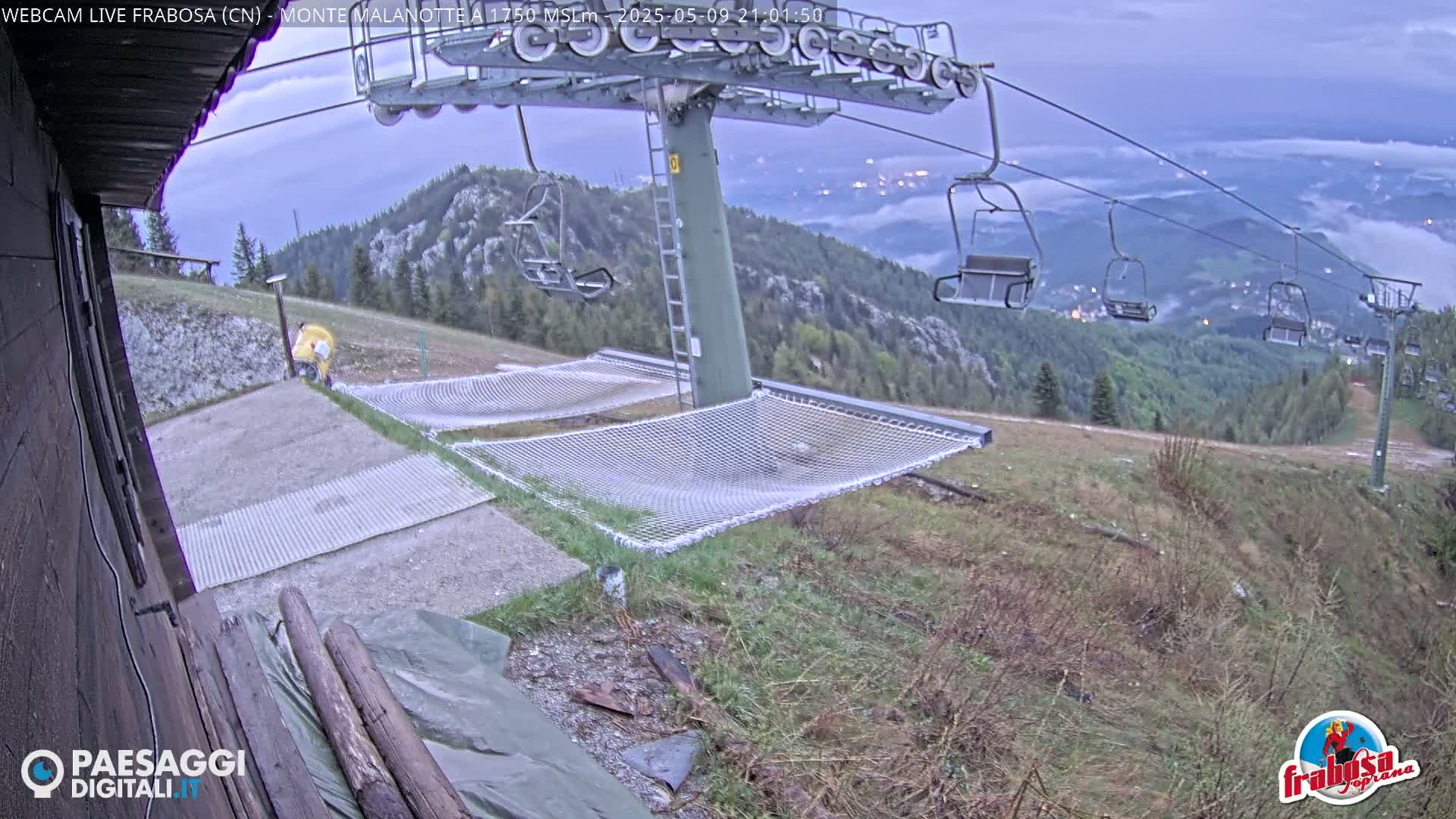 A chairlift and empty netting structures are situated on a grassy mountainside overlooking a valley shrouded in fog under a cloudy, twilight sky.
