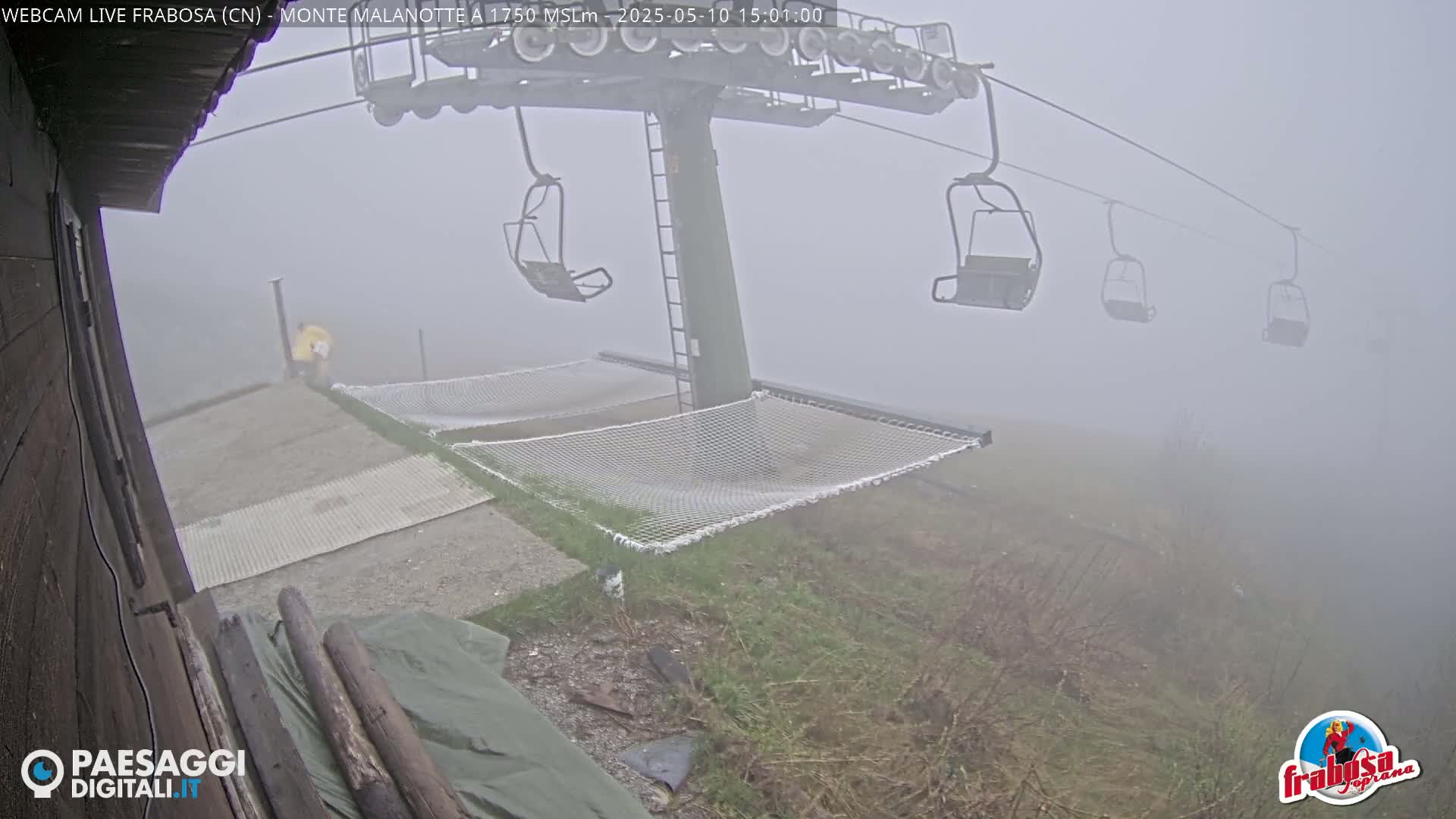 A person in yellow clothing stands near a foggy ski lift unloading station with several empty chairlifts and mesh hammocks.