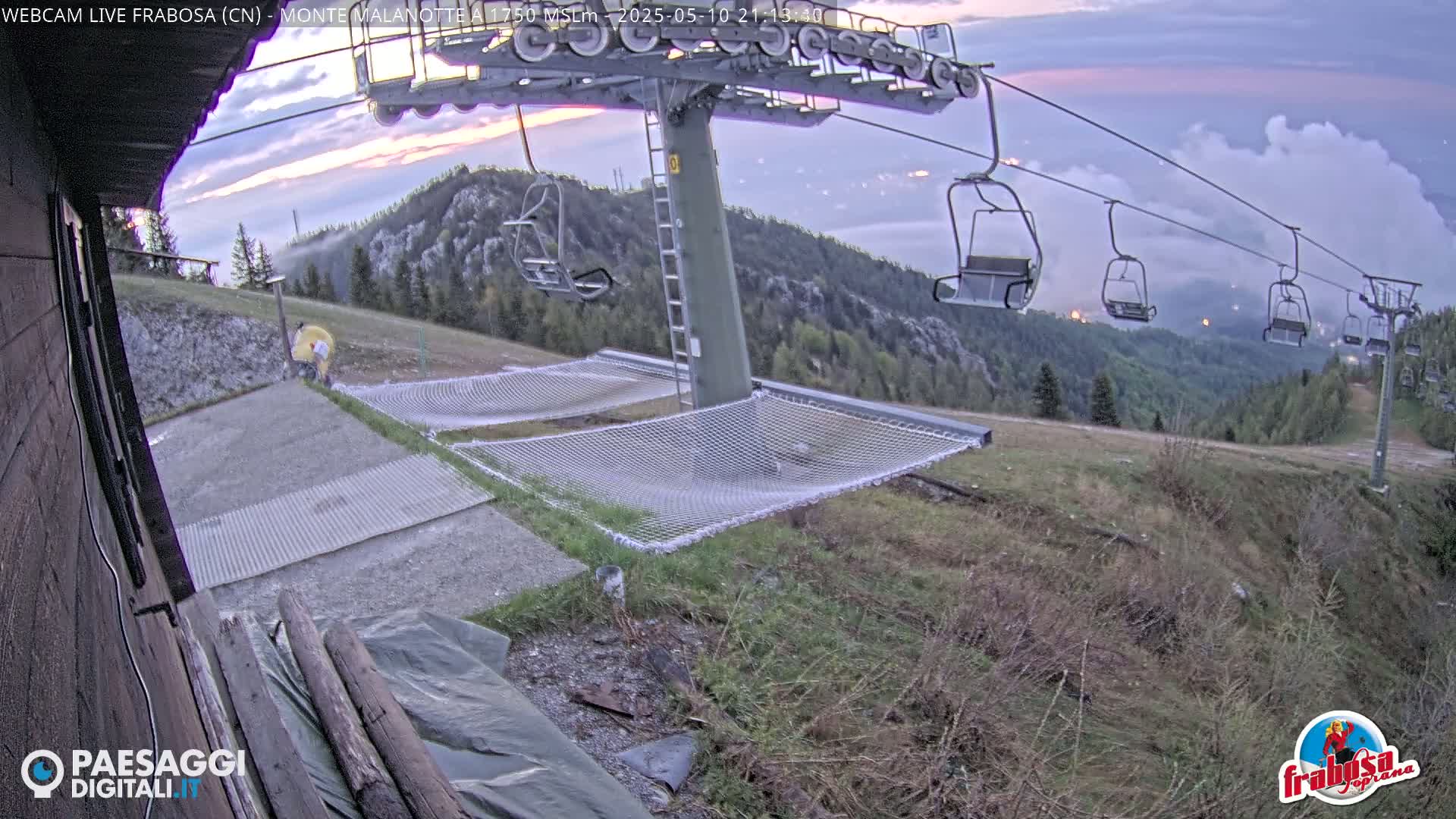 A chairlift extends across a mountaintop at dusk, with several empty chairs visible against a backdrop of low clouds and distant lights;  two nets are spread out below, and a grassy area leads down the slope.