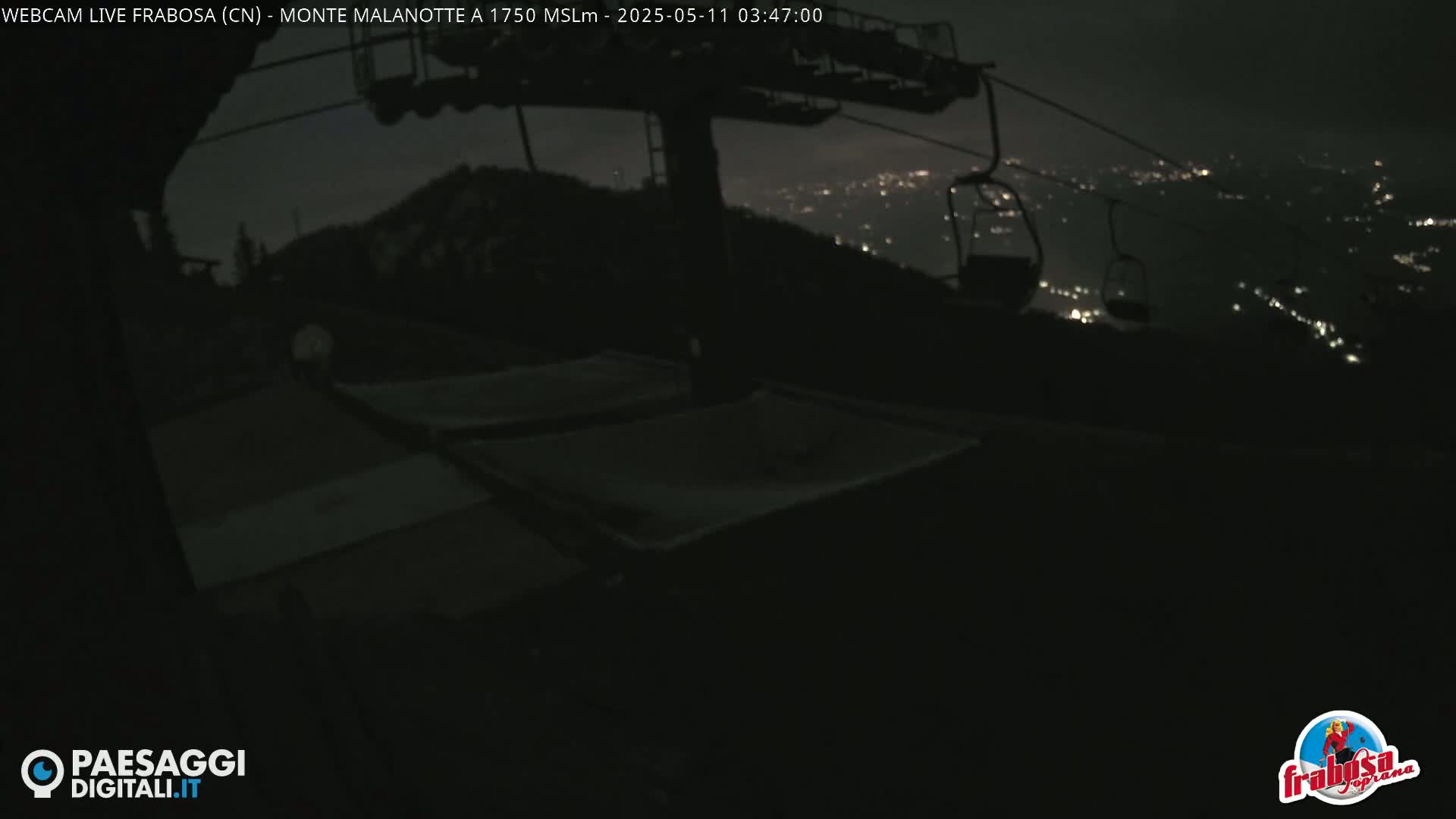 At night, empty chairlifts are visible against a backdrop of a distant, city's lights.