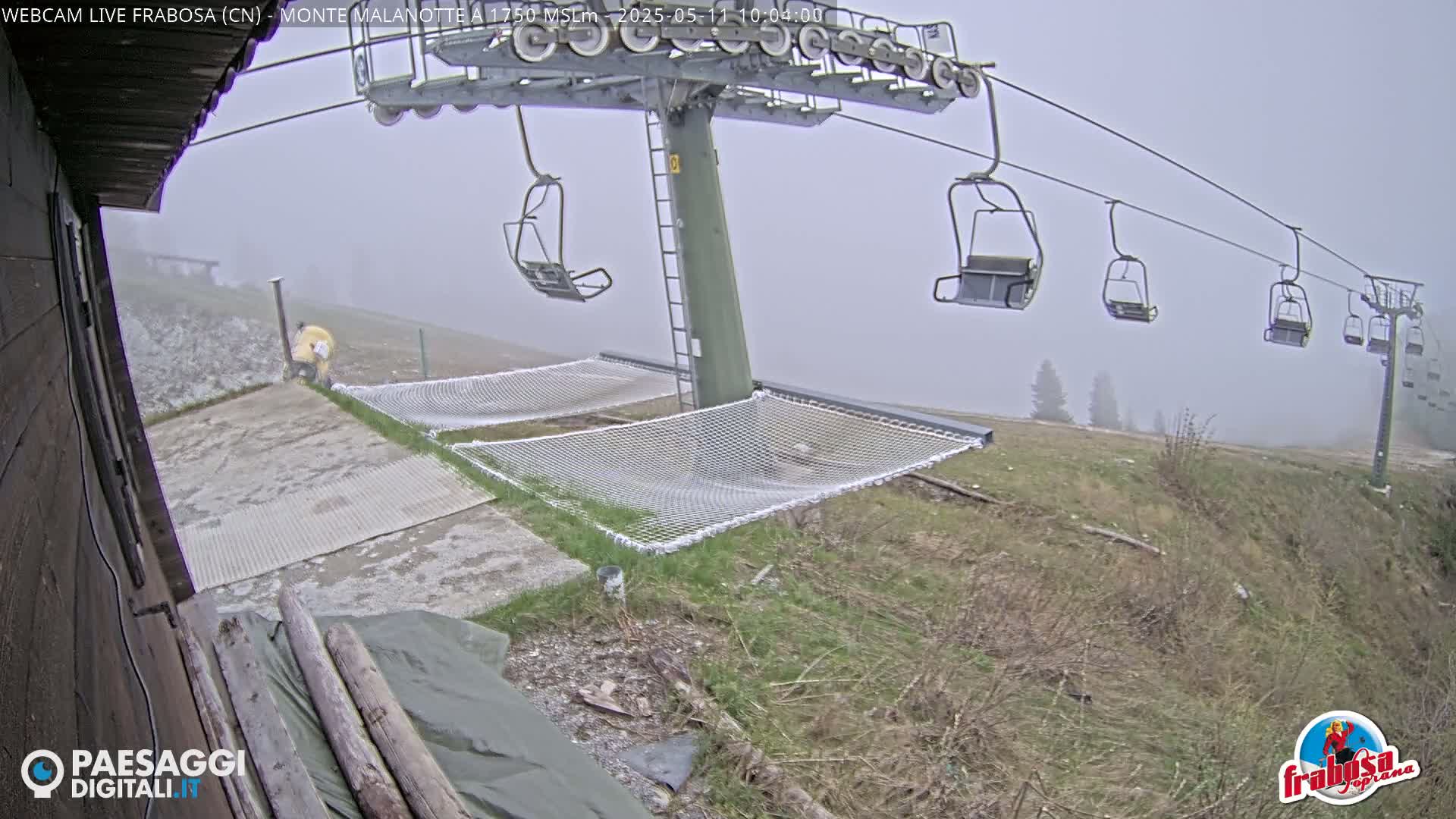 A foggy mountaintop view shows a chairlift with several empty chairs, safety nets, and sparse vegetation.