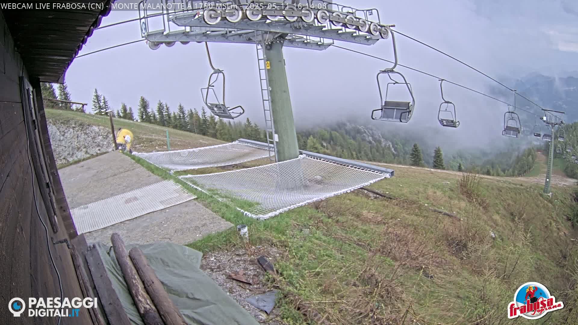 A chairlift with empty chairs ascends a foggy mountainside, passing over two empty mesh hammocks on the ground.