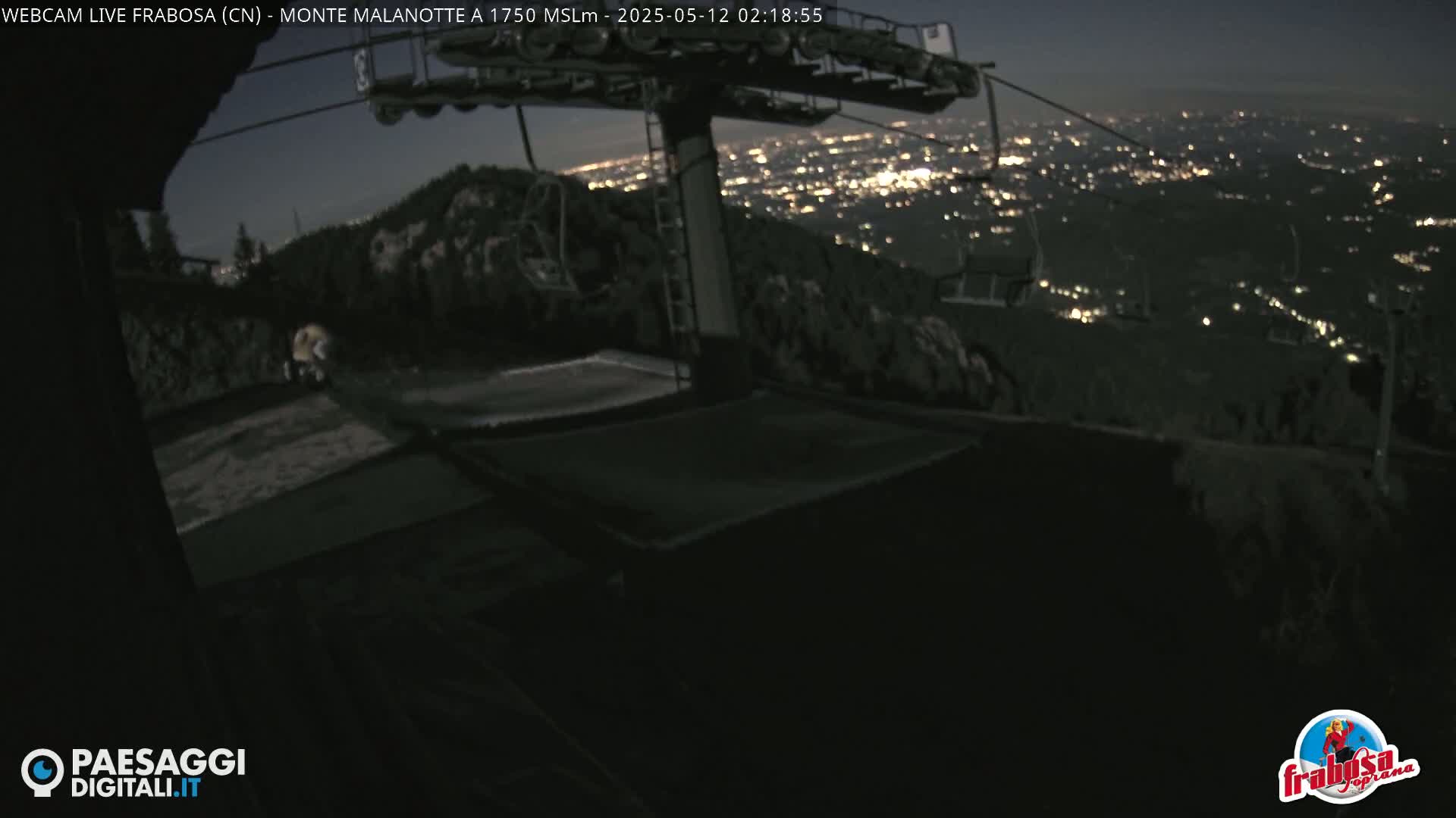 A nighttime view from a mountaintop shows a chairlift, a dark landscape, and a distant city glowing with lights.