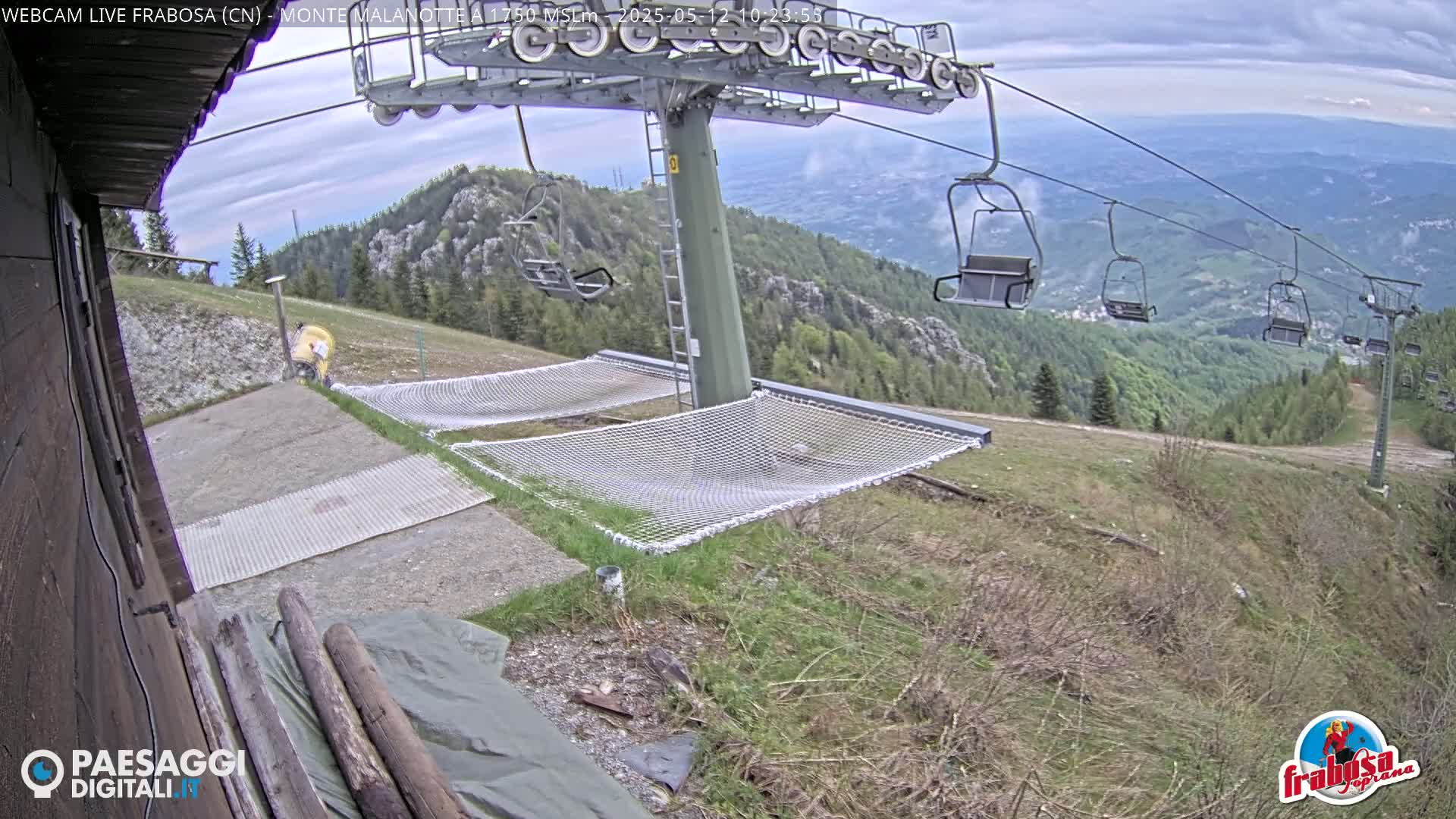 A mountaintop view shows an empty chairlift with several chairs visible, two empty mesh hammocks near a building, and an overcast sky.