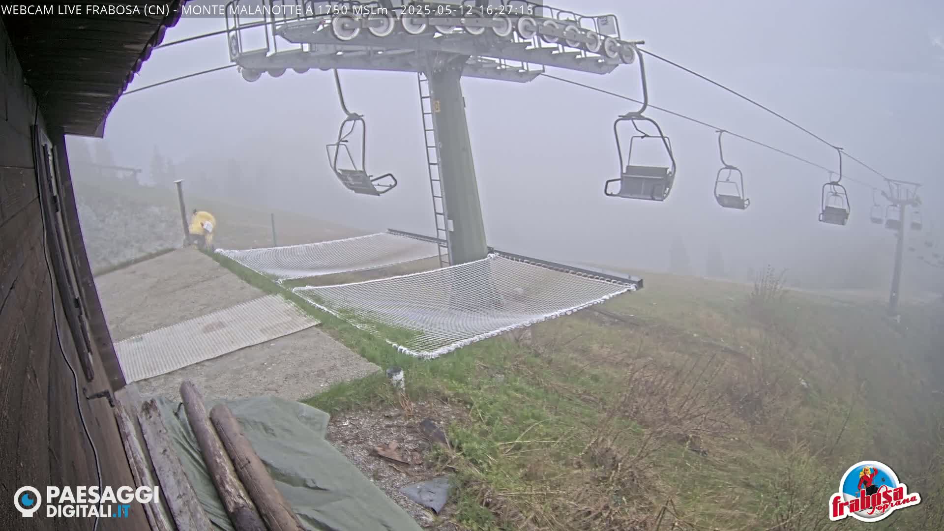 A foggy mountain scene shows a chairlift with empty chairs, two empty nets laid out on the ground below, and a person in a yellow jacket near the base of the lift.