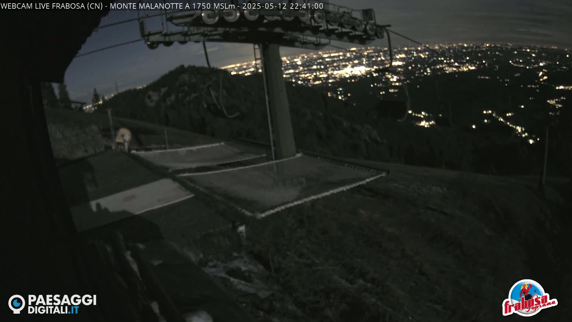 At night, a ski lift stands atop a mountain overlooking a city glowing with lights.