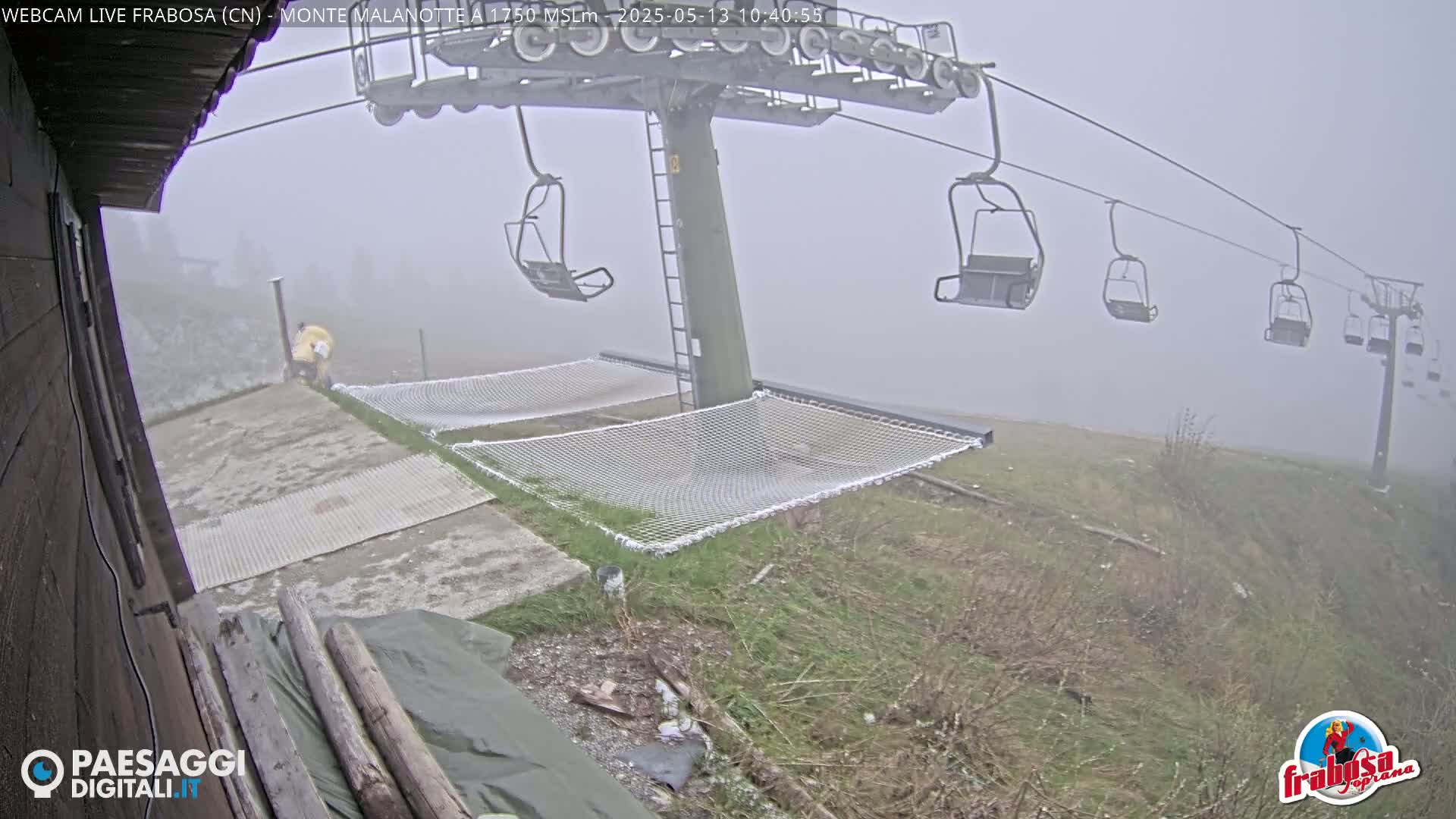 A foggy scene shows a chairlift with empty chairs and safety nets near a person in yellow clothing, all at the base of a mountain.