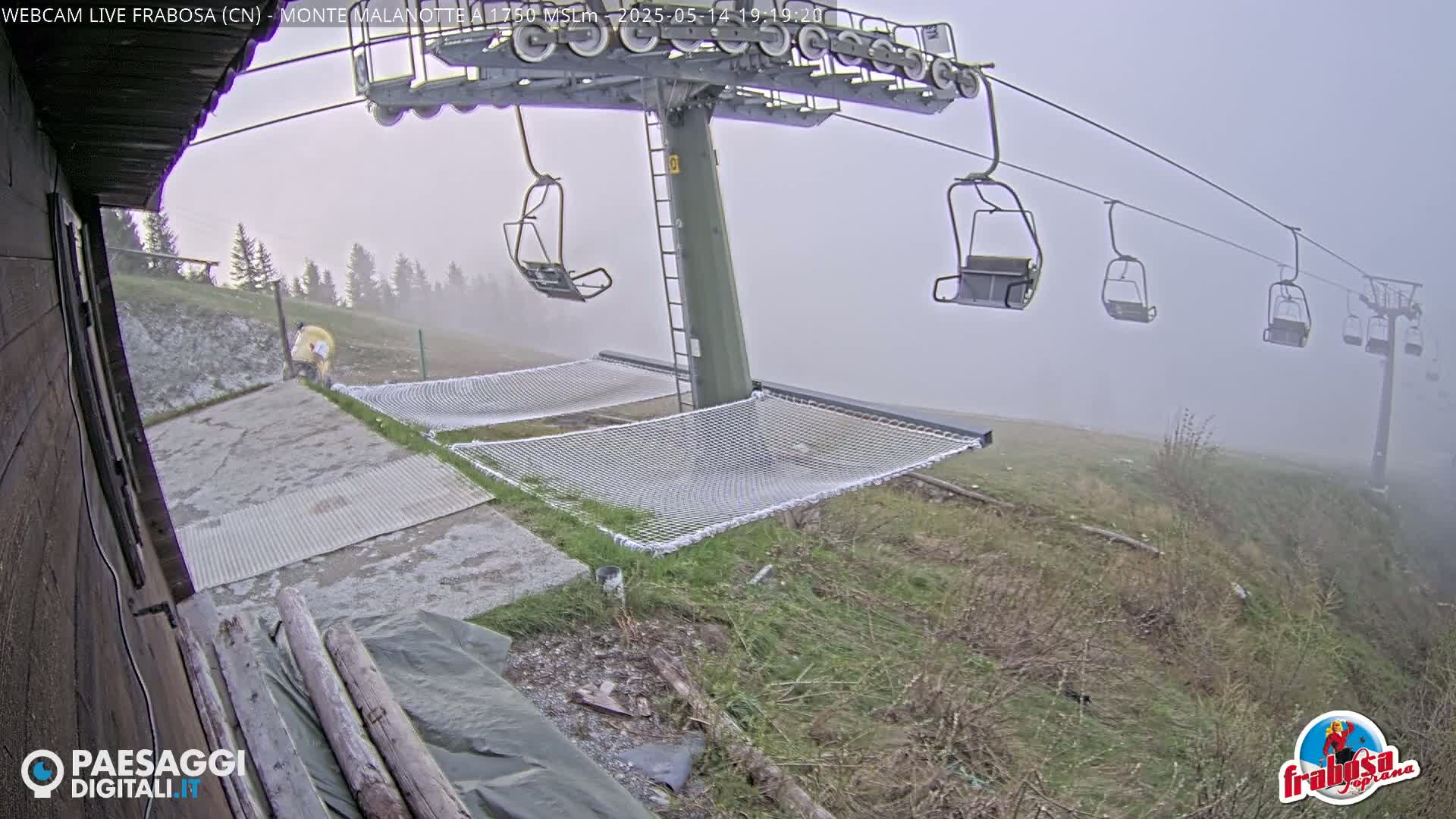 A foggy mountaintop scene shows a chairlift with empty chairs, two mesh hammocks, and a person in yellow clothing near a building.