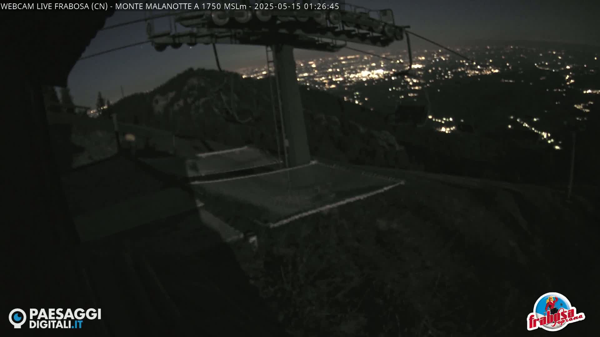 A nighttime view from a mountaintop shows a chairlift and a distant city illuminated below.