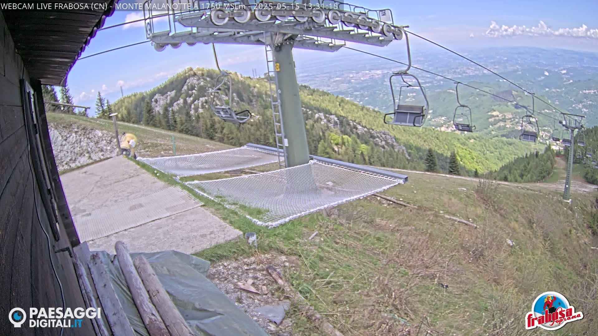 A mostly sunny mountaintop scene shows a chairlift, several empty chairlift seats, and two net hammocks near a building, with a person visible in the distance.