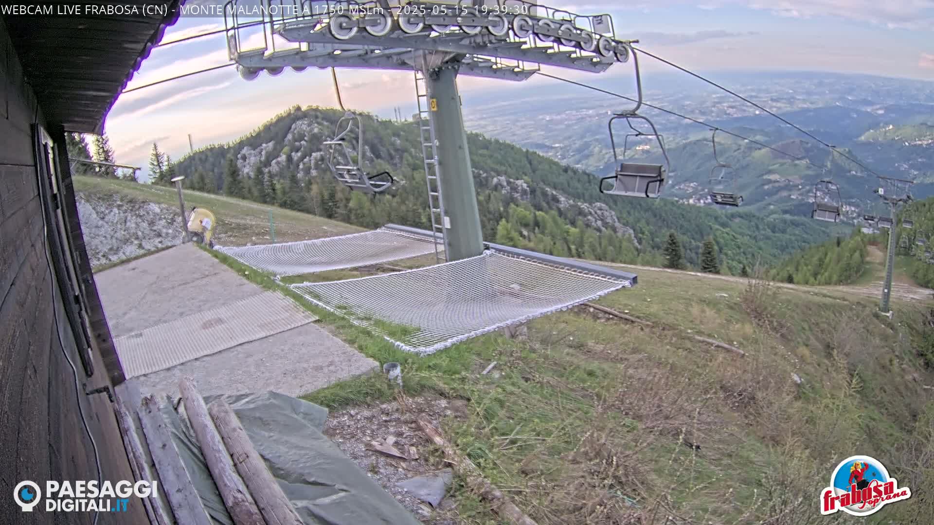 A mountaintop ski lift with empty chairs overlooks a valley under partly cloudy skies, with two net hammocks situated near a small building.