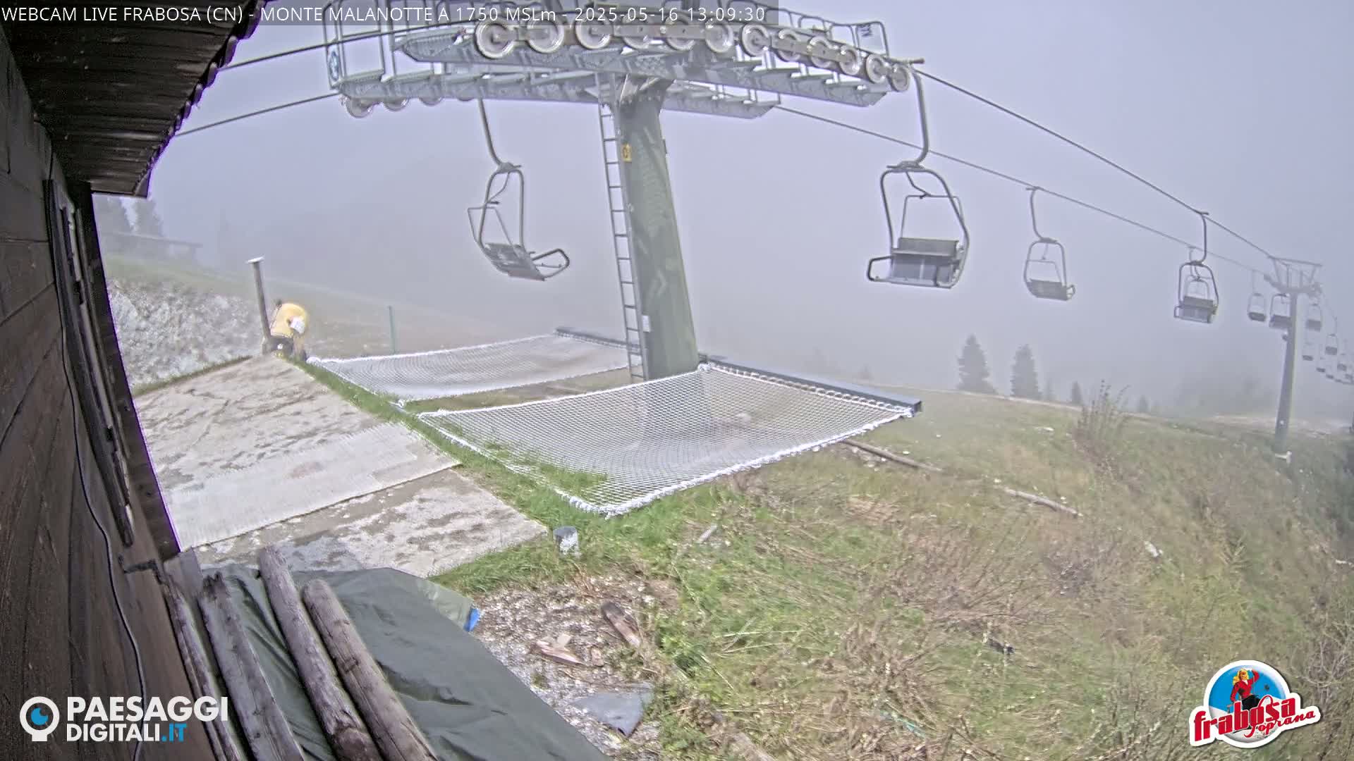 A foggy mountaintop scene shows a chairlift with empty chairs and several netting hammocks near a building, with a person in yellow clothing visible in the foreground.